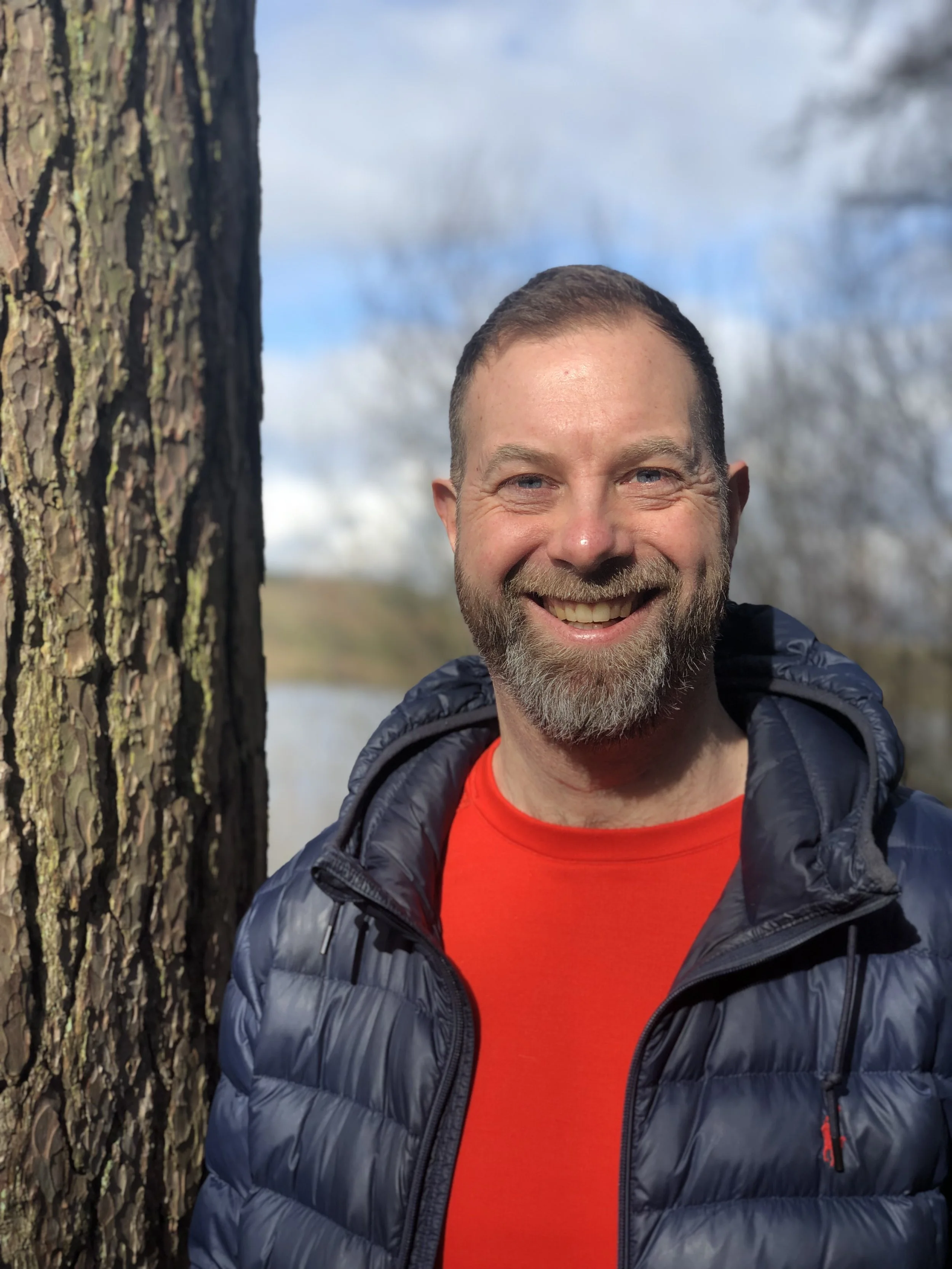 Smiling man with beard in outdoor setting near a tree, wearing a navy puffer jacket and red shirt, with a background of water and cloudy sky.