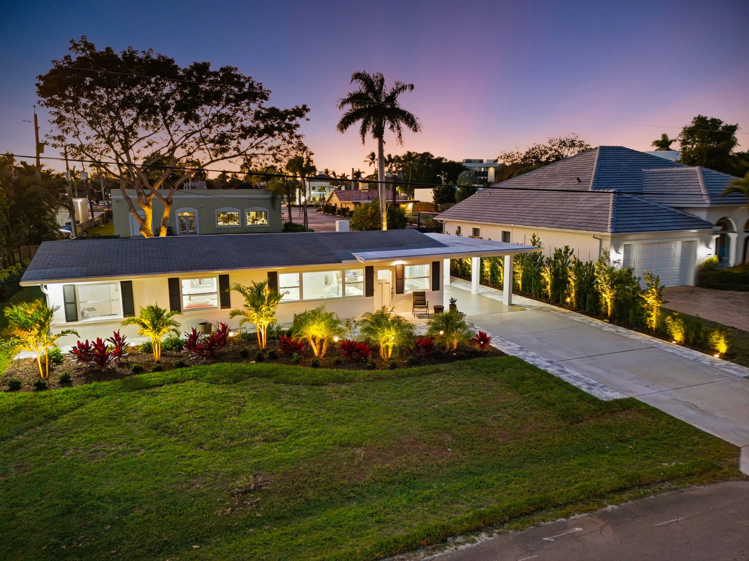 A modern house at dusk with well-lit exterior, front yard with landscaped plants, a driveway, and neighboring houses in the background.