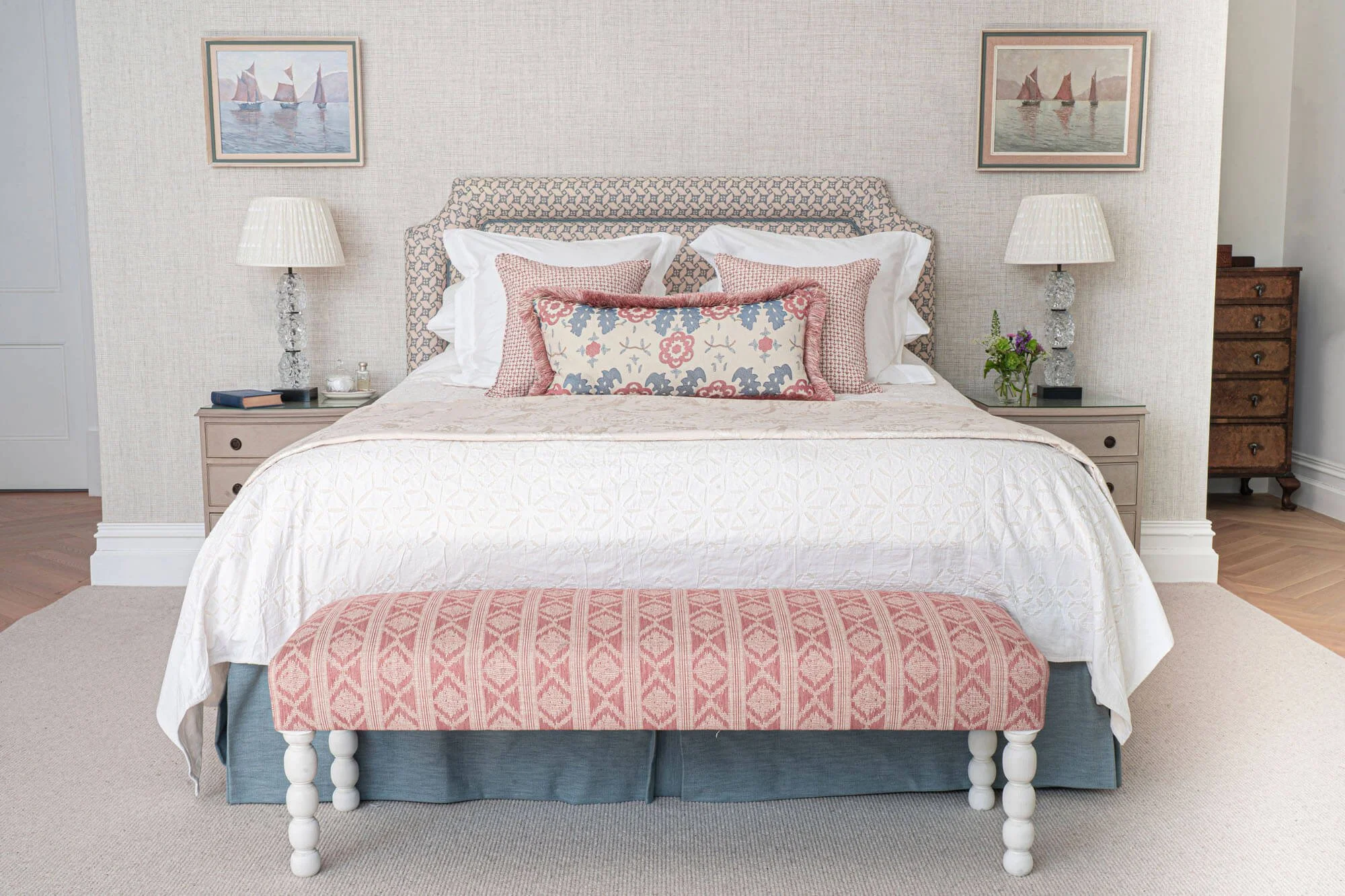 A neatly made bed with multiple pillows and a patterned headboard, flanked by two bedside tables with lamps, in a stylish bedroom with framed nautical art on the wall.