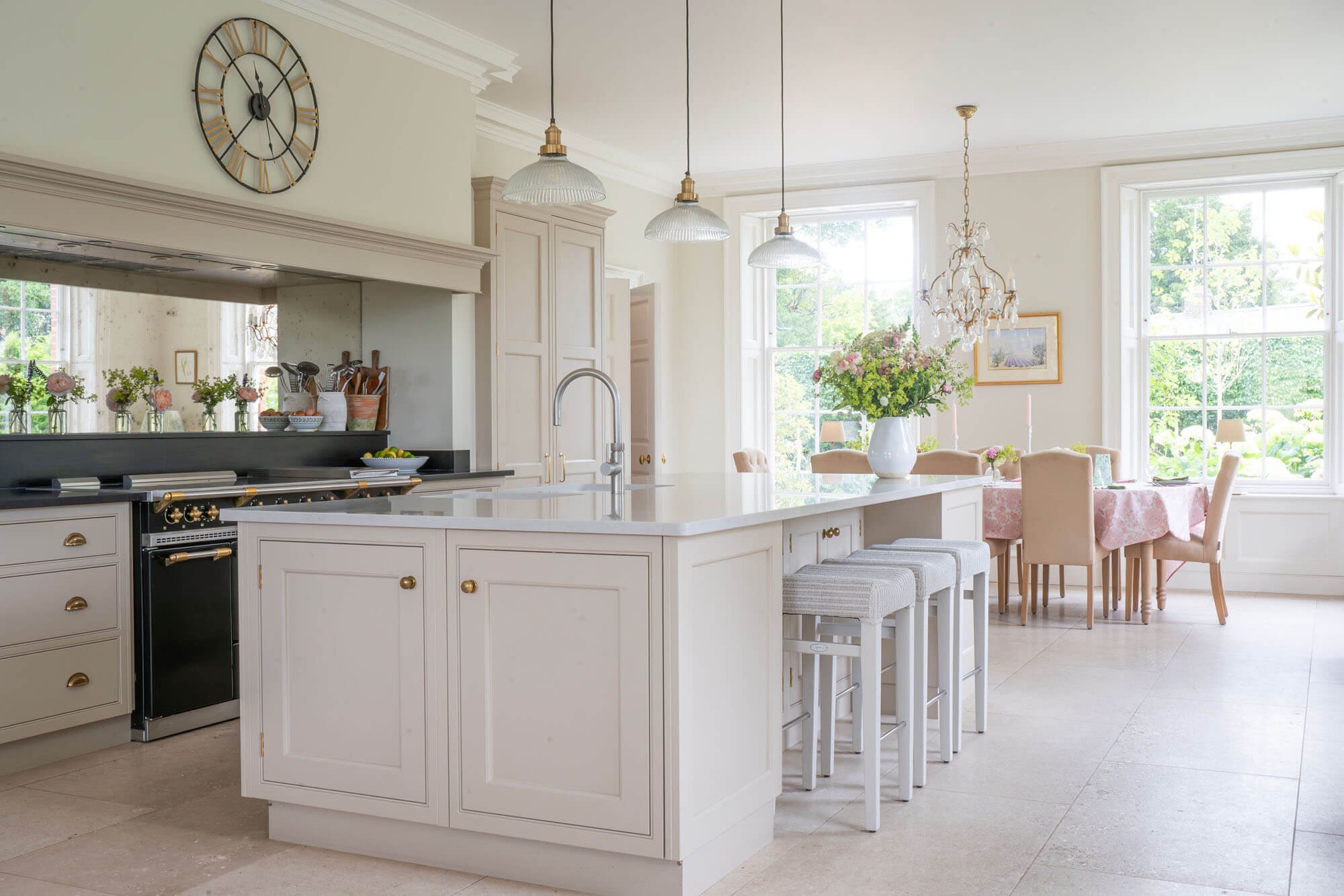 Bright, airy kitchen and dining area with white cabinets, large windows, a central island with bar stools, and a dining table with chairs and a vase of flowers.