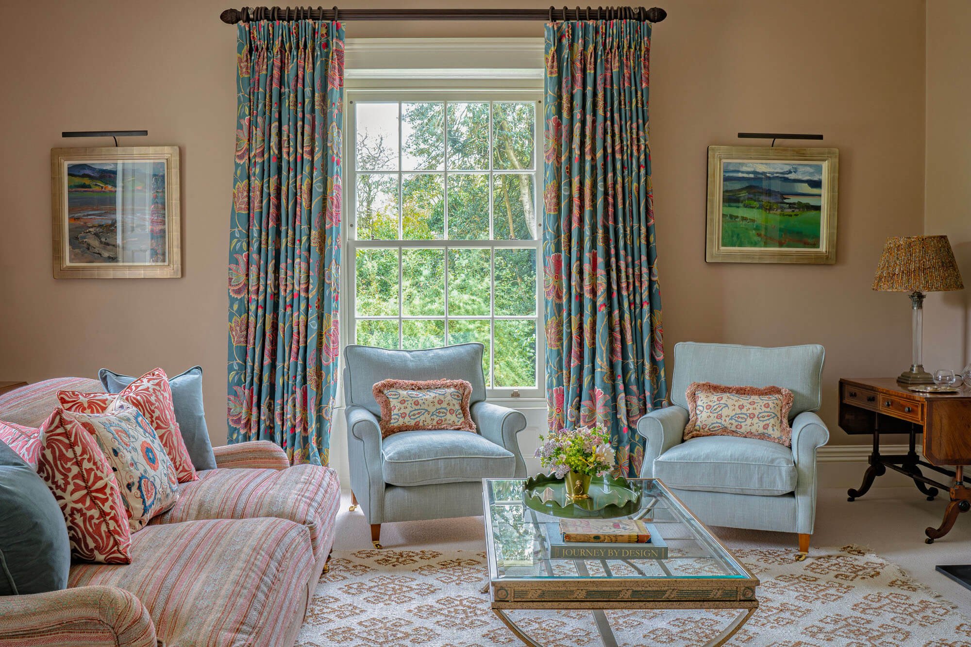 A cozy living room with a large window, blue upholstered armchairs, a pink striped sofa with colorful pillows, a glass coffee table with books and flowers, and framed landscape art on beige walls.