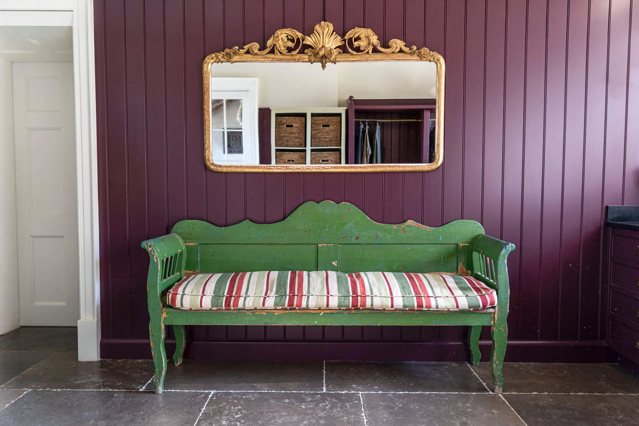 A vintage green wooden bench with a striped cushion in front of a purple paneled wall. A gold ornate mirror hangs above the bench, reflecting a closet with shelves and baskets.