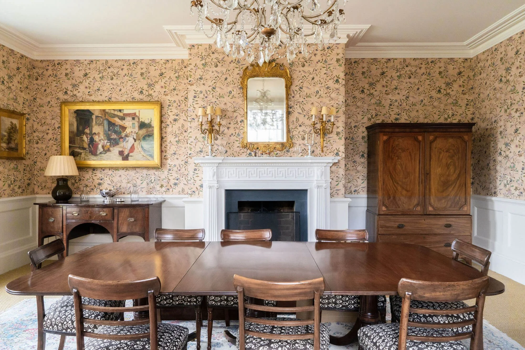 A formal dining room with vintage decor, a wooden table with eight chairs, a white fireplace mantel with a mirror, floral wallpaper, a chandelier, a painting, a sideboard with a lamp, a wooden cabinet, and decorative elements.