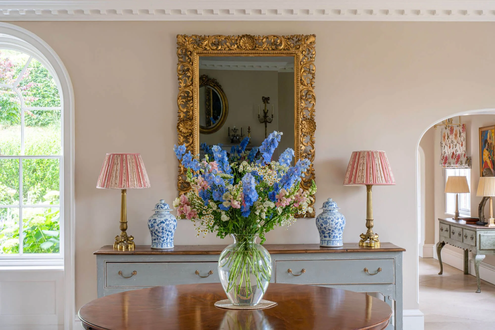 A vase of blue, pink, and white flowers on a polished wooden table in a well-lit room with a large window, a gold-framed mirror, and two matching table lamps with pink striped shades on a light-colored console table.