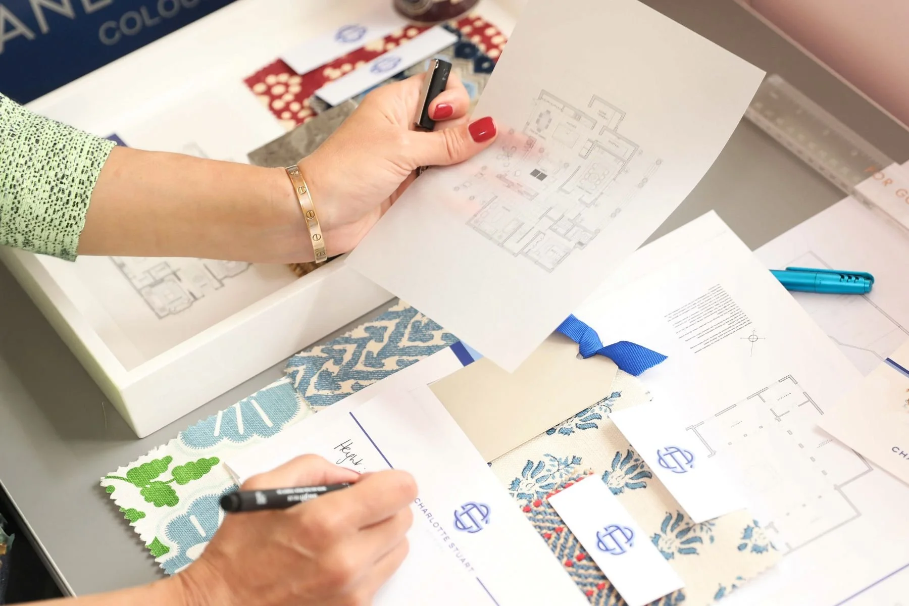 Person holding a printed house plans and a pen, working on interior design or home blueprint at a table with fabric samples and additional architectural drawings.