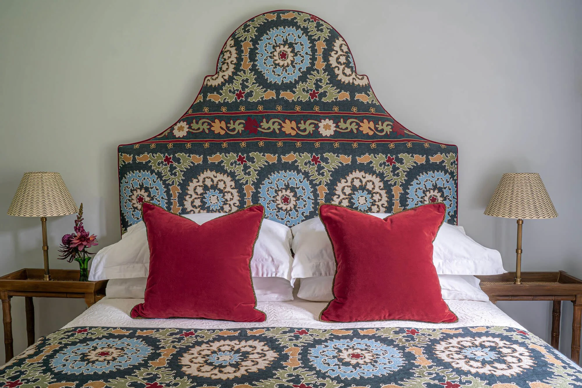 A bed with a patterned headboard, two white pillows, two red velvet pillows, and a matching bedspread. Two bedside tables with lamps and a small flower arrangement are on either side of the bed.