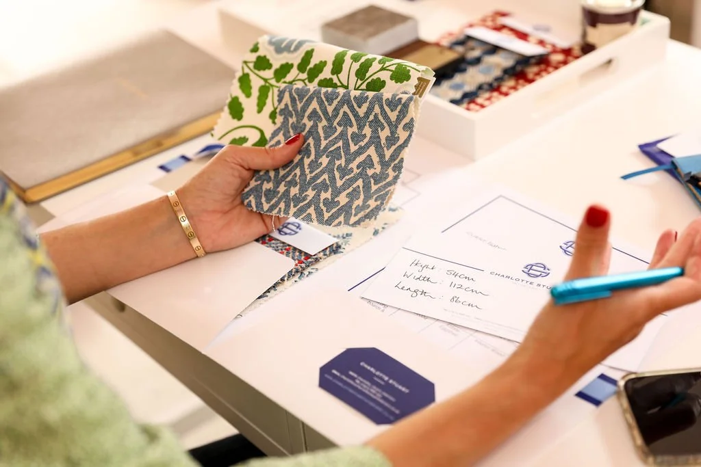 Person holding fabric samples while sitting at a table with papers, pens, and a box of fabric swatches.
