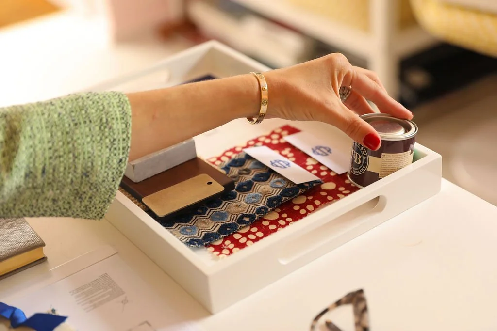 A woman reaching into a white tray containing fabric swatches, a small jar, and a beige card, on a white surface.