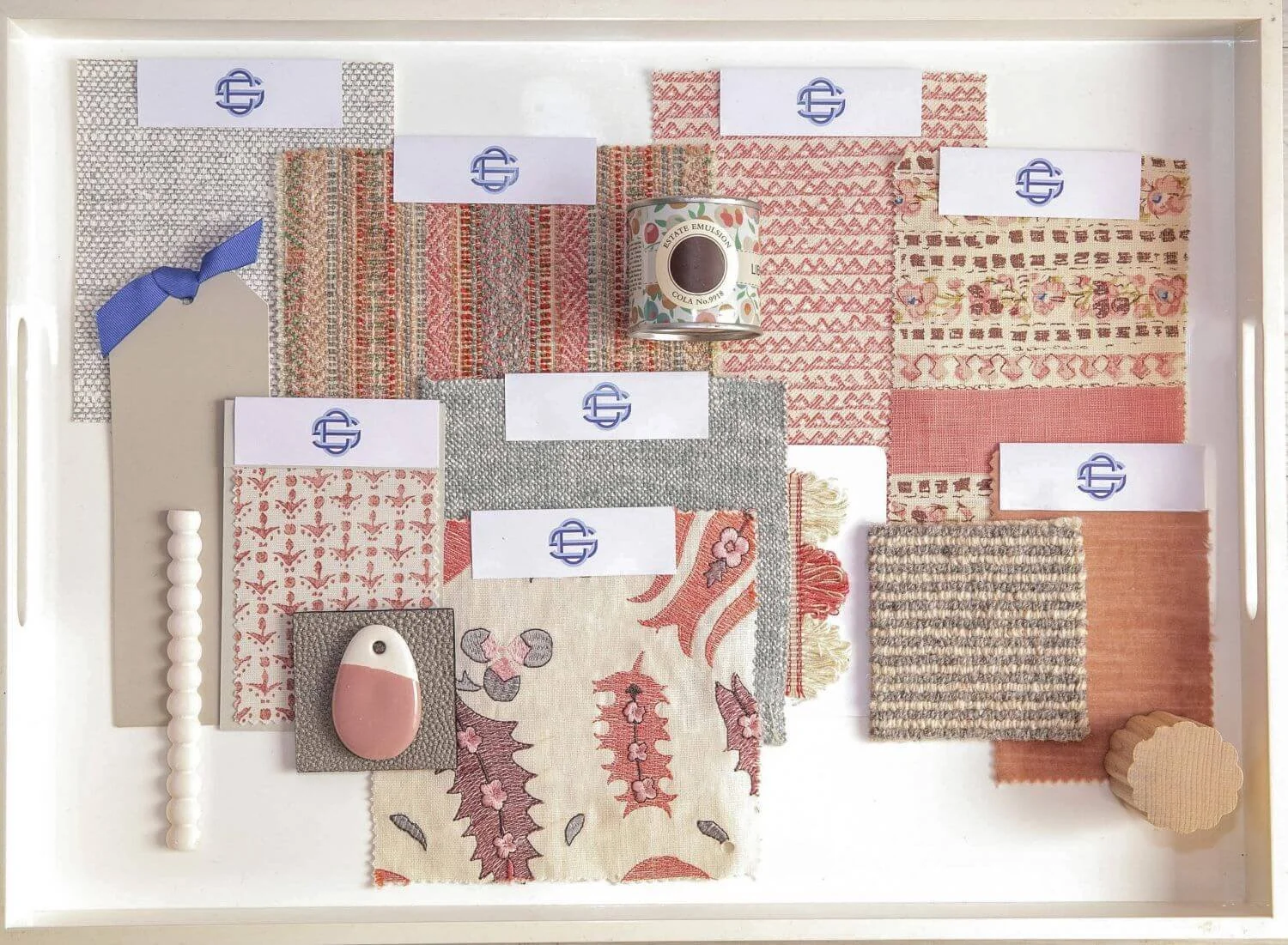 A display of fabric swatches in various patterns and colors, including shades of pink, red, gray, and beige, arranged on a white tray with white labels featuring a blue logo. There is also a pink and beige pebble-shaped object, a small wooden spool, and a string of white beads on the tray.