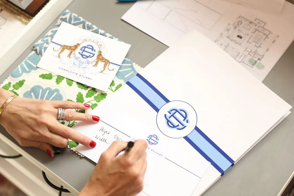 A person with red nail polish and rings writing on a sheet of paper with a marker, with a Charlotte Stuart branded folder and business card on a desk, along with architectural plans and a printed floor plan in the background.