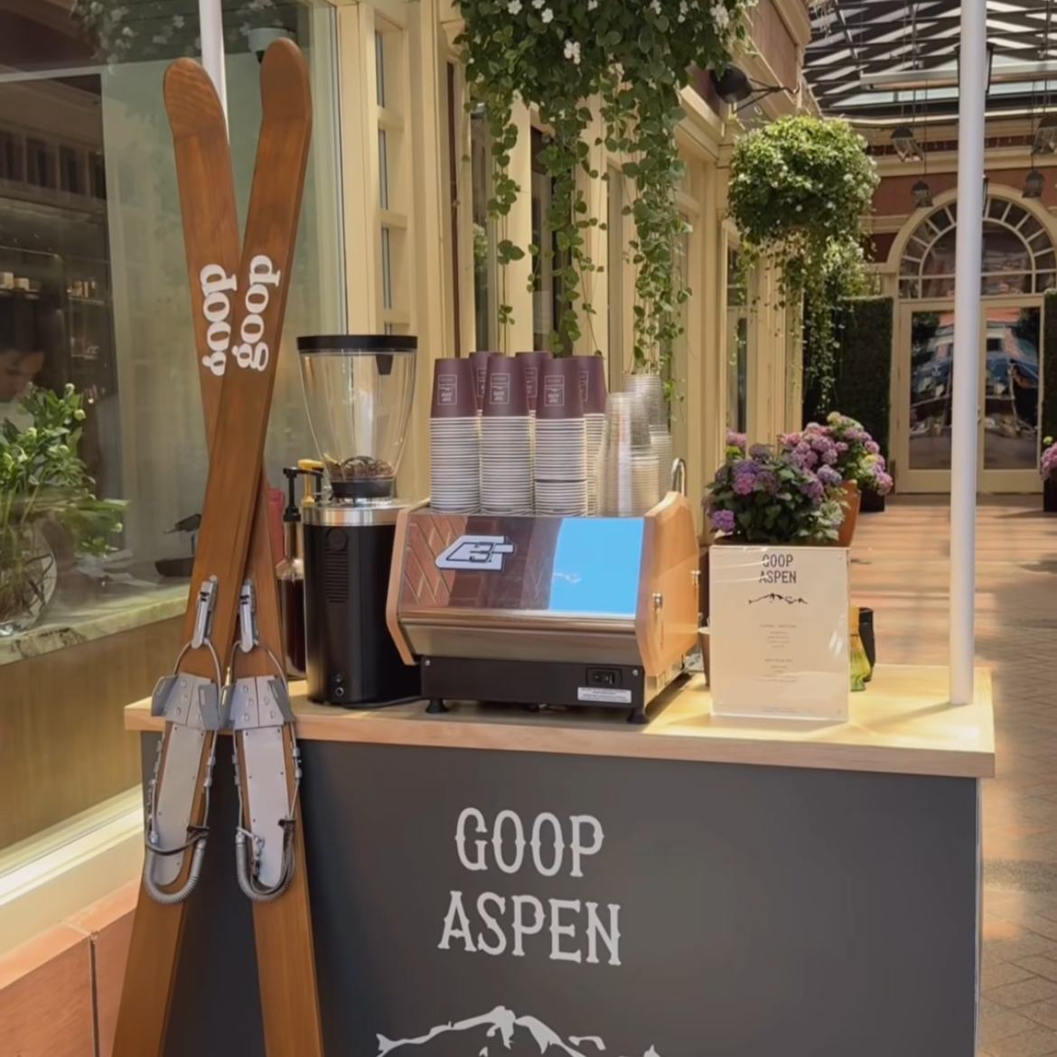 custom coffee cart catering station with cups, a coffee grinder, and a sign that says 'Goop Aspen.' Ski poles leaning against the side and surrounded by flowers and hanging plants.