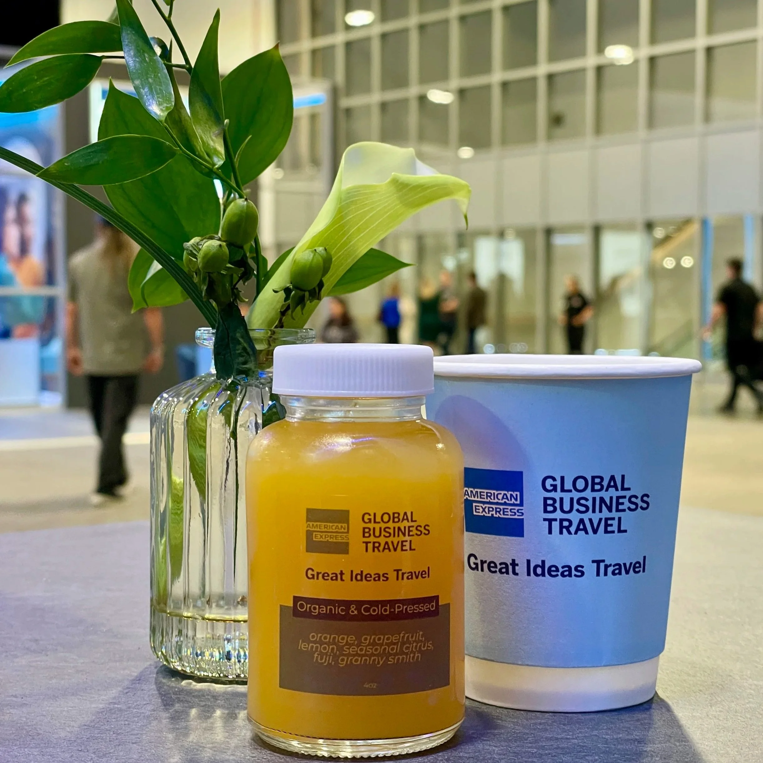 A yellow bottle of organic cold-pressed juice with a white cap, a clear glass vase with green leaves and a calla lily, and a white cup with blue text, all on a table inside a modern building with large glass windows and people in the background