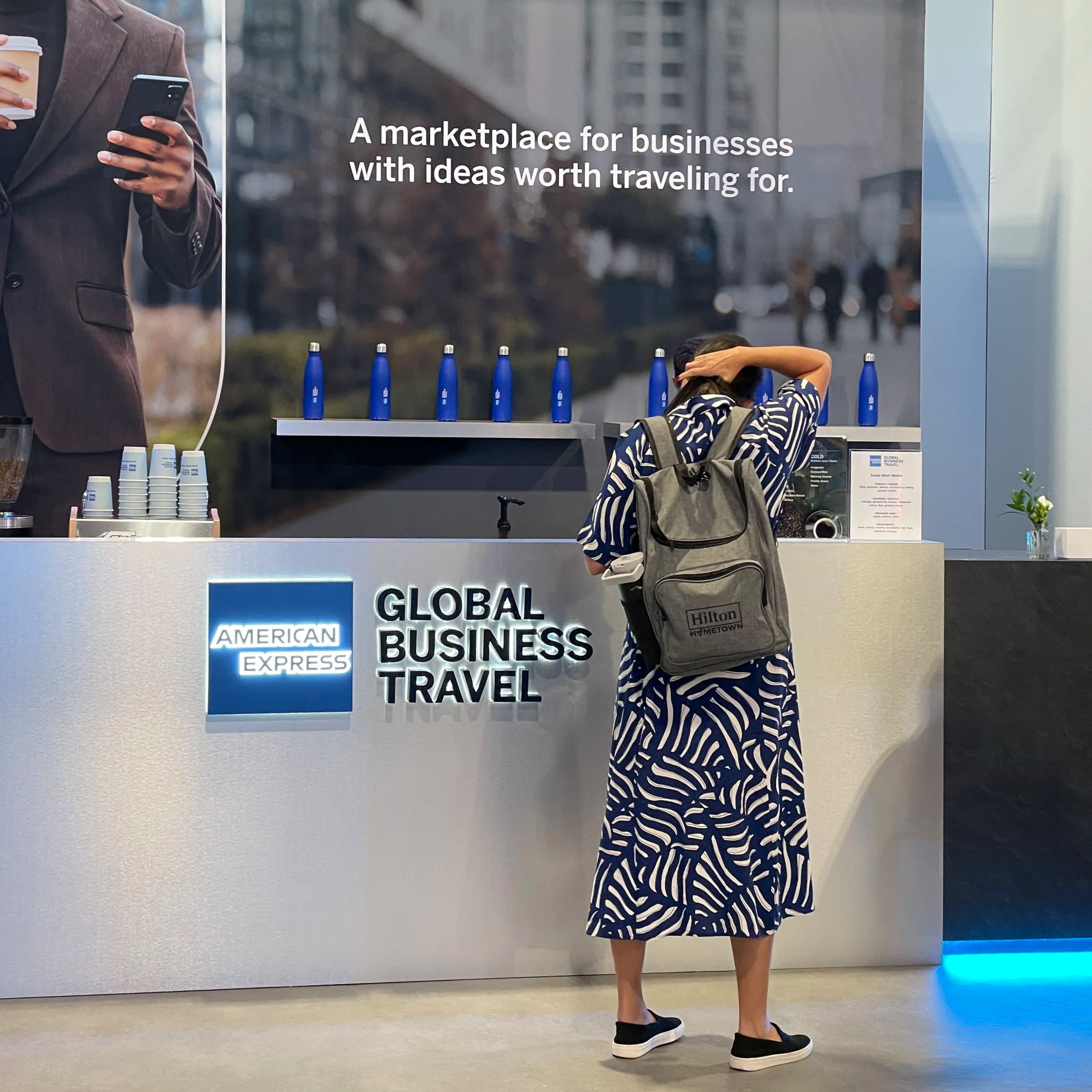 A woman with a backpack standing at an American Express Global Business Travel espresso counter, talking to barista, in a modern expo at colorado convention center coffee cart catering