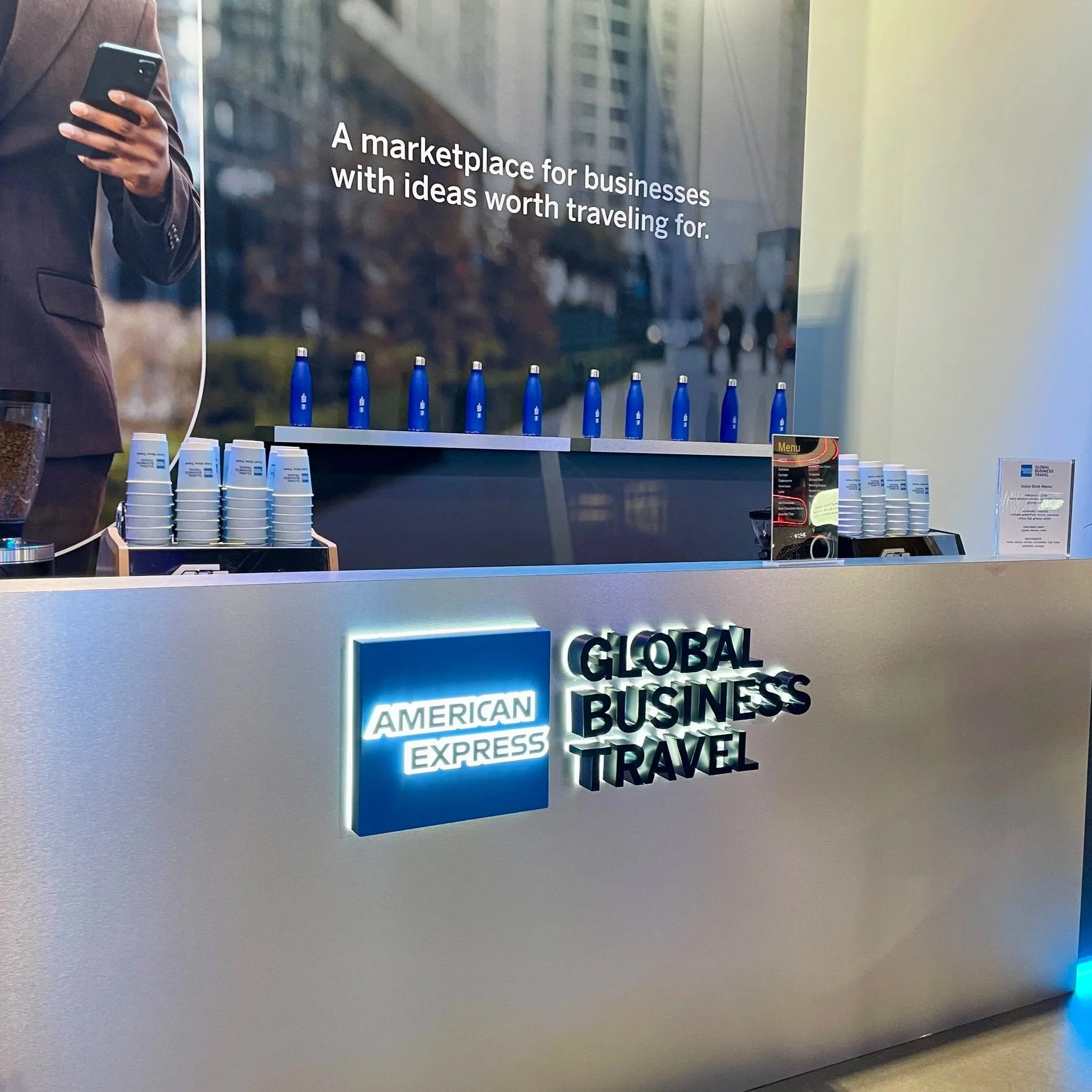 American Express logo on a sign with the words 'Global Business Travel' at a service counter in an airport or travel agency. A person in a suit is seen holding a phone in the background, with a large advertisement or backdrop behind the counter.
