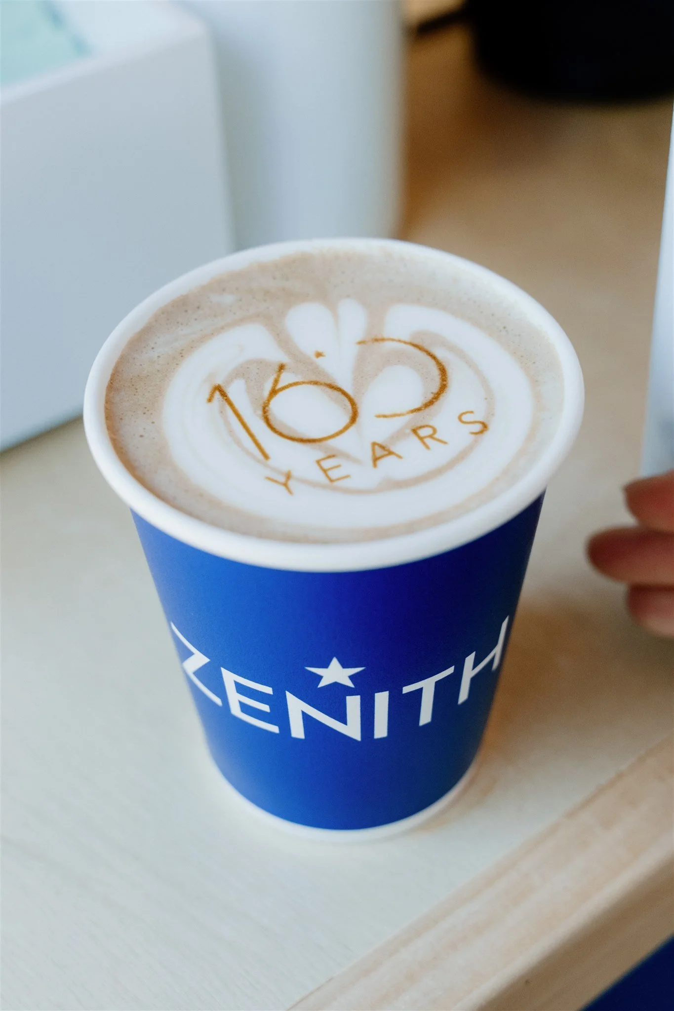 A cup of coffee with latte art that says '1823 YEARS' inside a circle, in a blue cup with 'ZENITH' written on it.