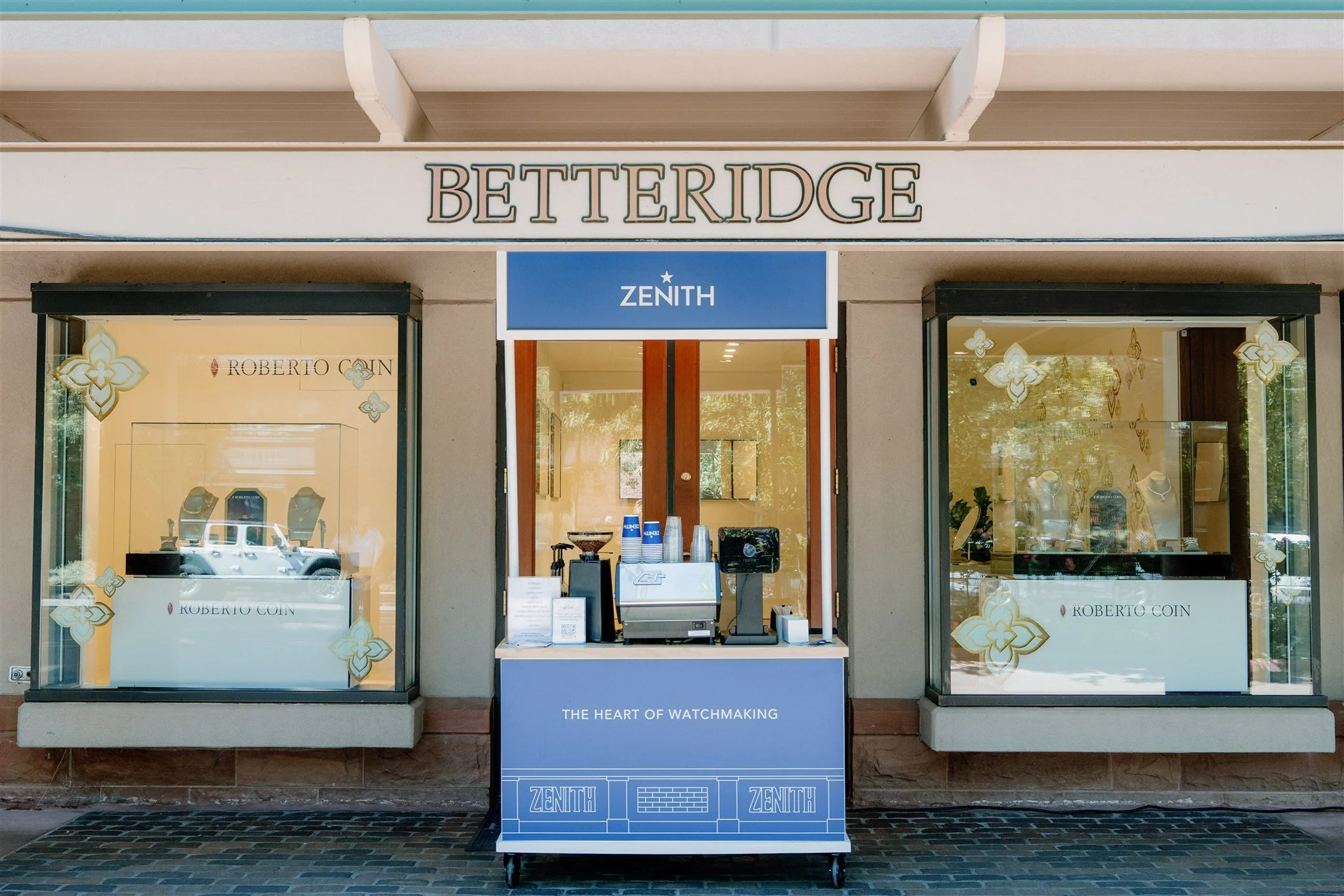 custom coffee cart brand activation of a watch store named BETTERIDGE with a blue stand in front labeled ZENITH, featuring coffee and supplies, flanked by two windows showcasing watches and jewelry.