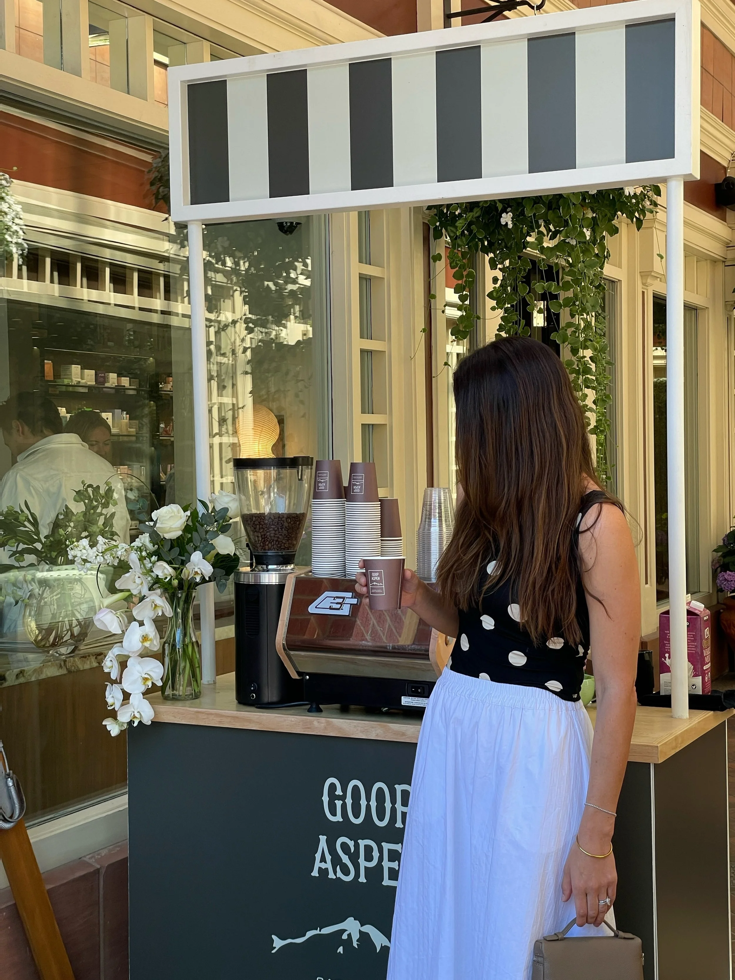 A woman ordering coffee at a branded café stand labeled 'Good Aspen' with a flower arrangement nearby, coffee grinder, and cups on the counter.
