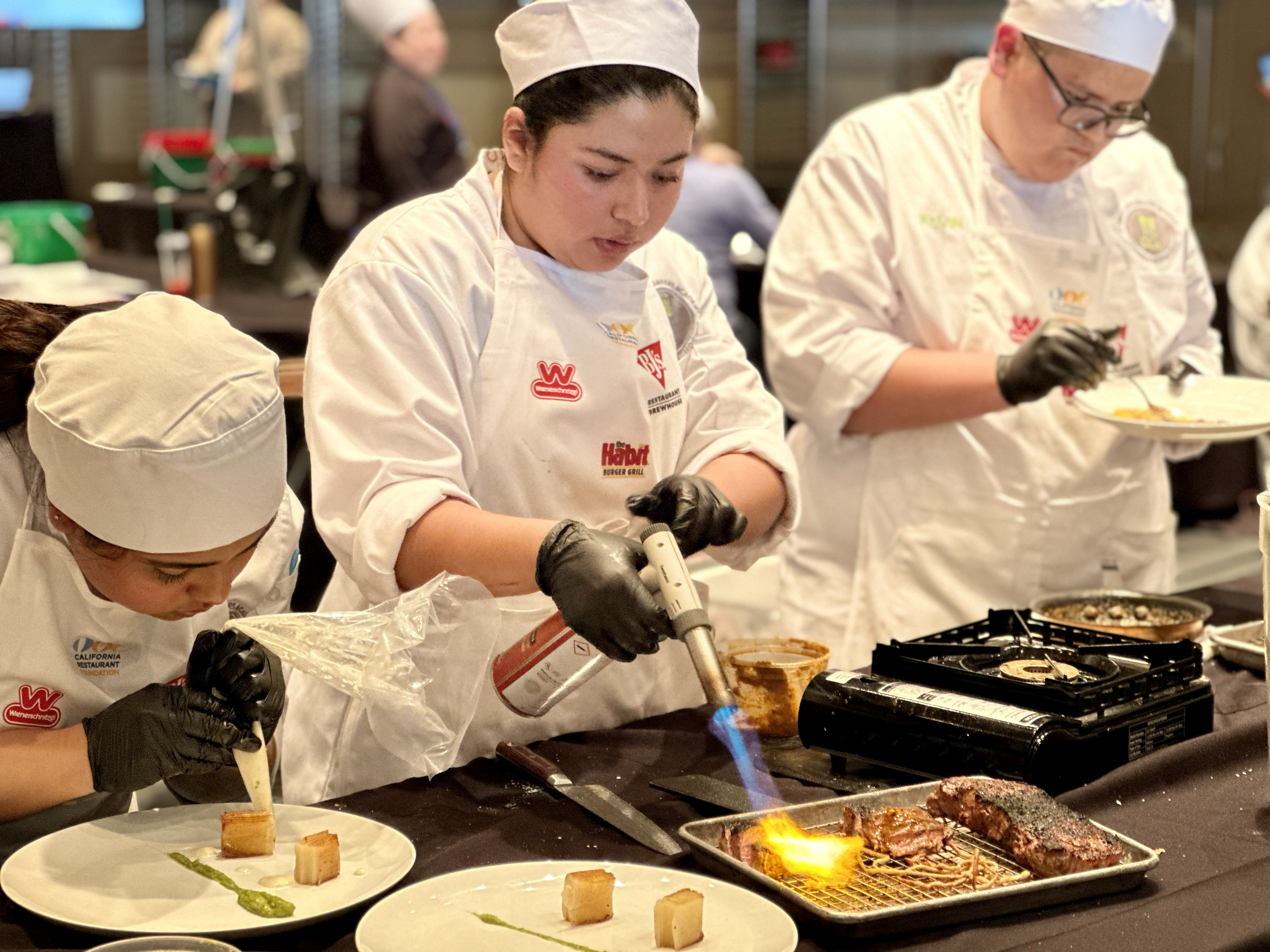 Chefs in white uniforms and hats cooking and plating food in a professional kitchen, with grilled meat on a tray and plates with small food portions.