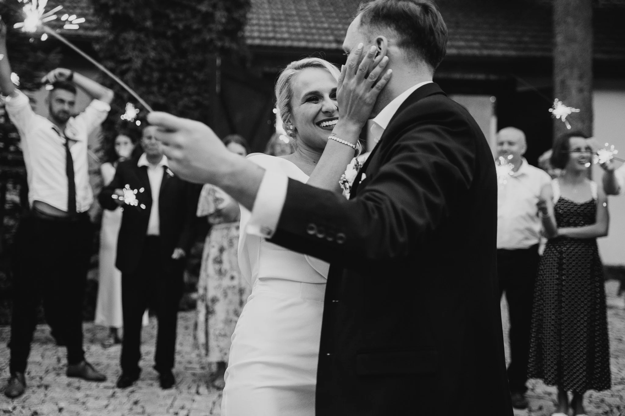 A black and white photo of a bride and groom dancing closely during their wedding celebration, surrounded by guests holding sparklers.