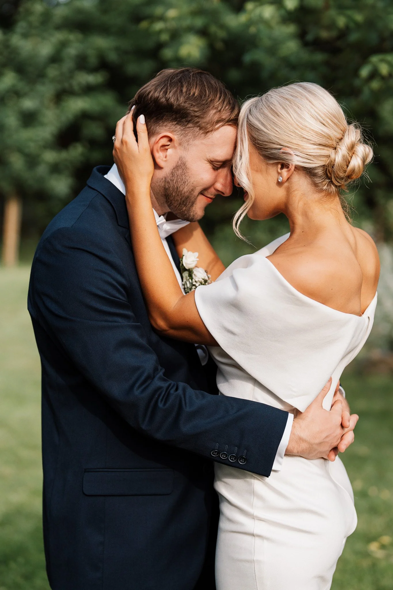 A couple dressed in wedding attire embracing outdoors, with their foreheads touching and eyes closed, in a green park setting.