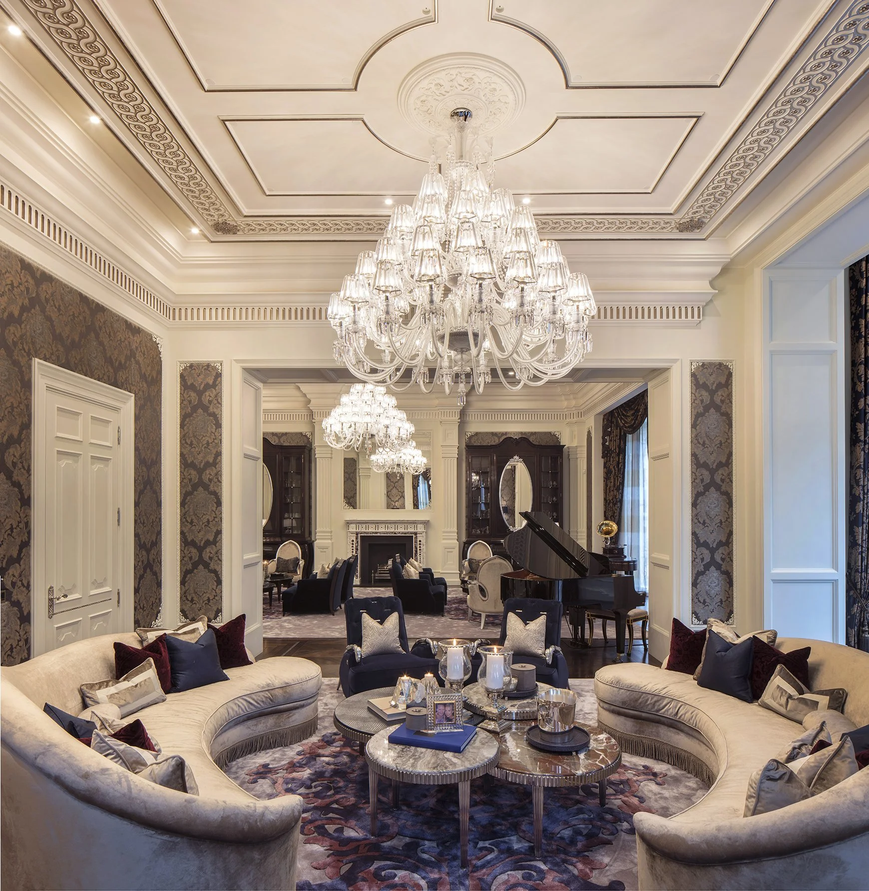 Elegant living room with ornate crown molding, a large crystal chandelier hanging from a decorative ceiling, and a mix of luxurious furniture including curved sofas, armchairs, and a grand piano. The room features patterned wallpaper, dark wood-panel
