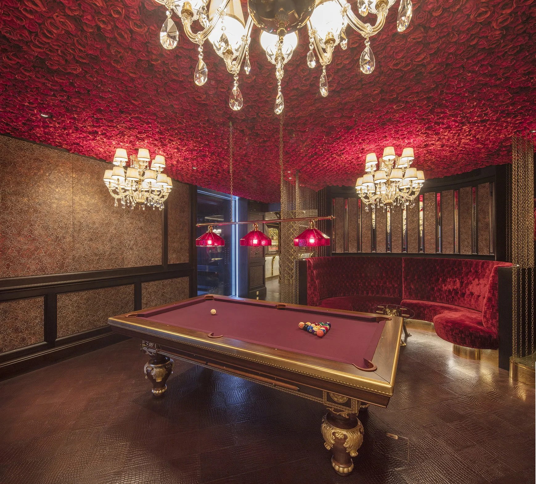 A luxurious interior featuring a maroon pool table with billiard balls racked, surrounded by ornate chandeliers and a ceiling covered in red roses. The room has dark wood paneling and elegant lighting fixtures.