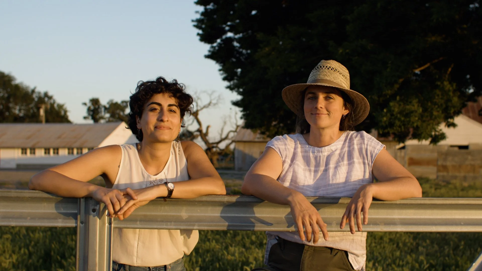 Two women leaning on a metal fence outdoors, smiling at the camera during golden hour, with trees and buildings in the background.