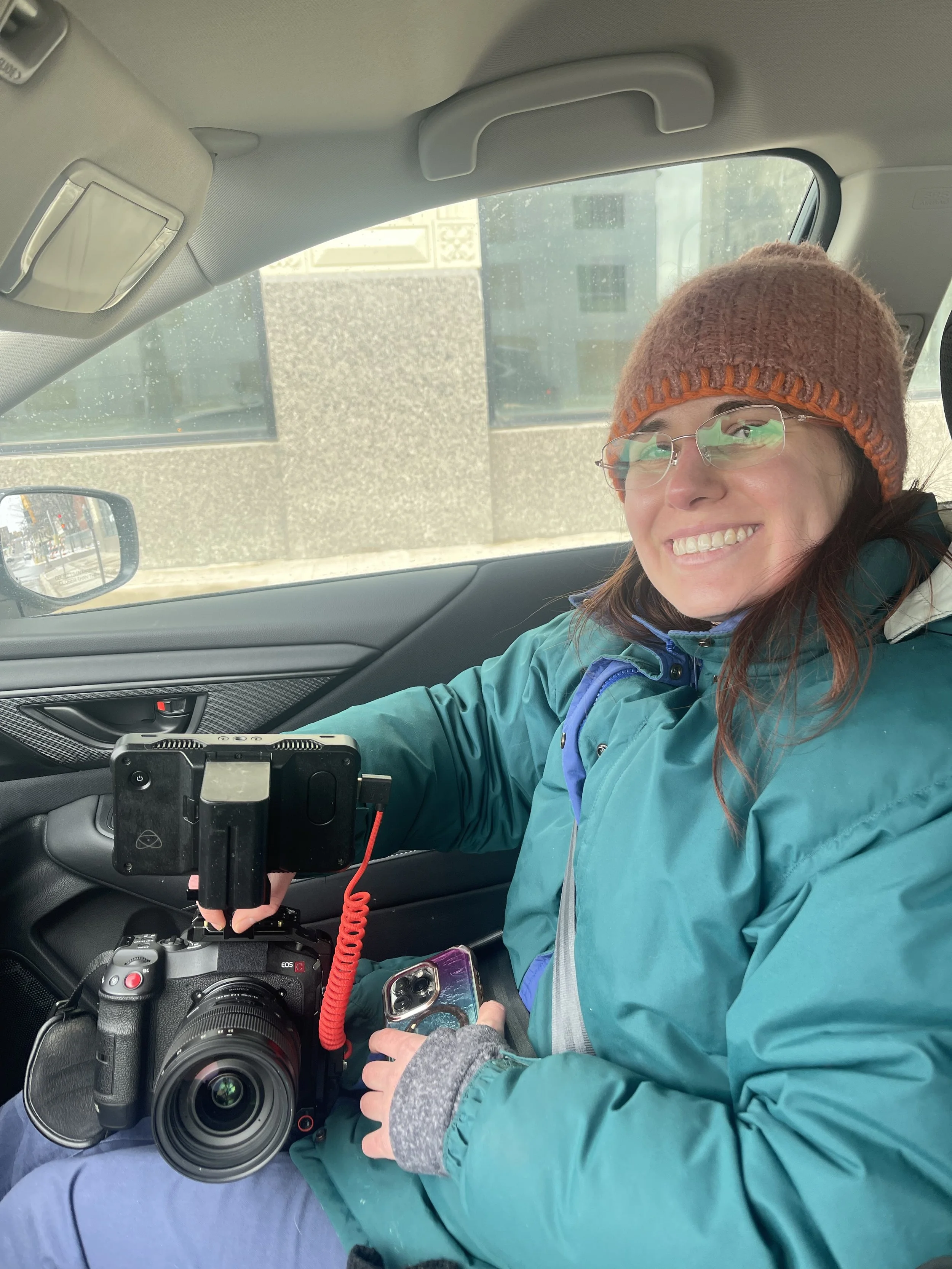Woman sitting in a car, smiling, wearing glasses, a brown knit hat, a teal jacket, holding a professional camera with a red strap, and a smartphone in her hand.