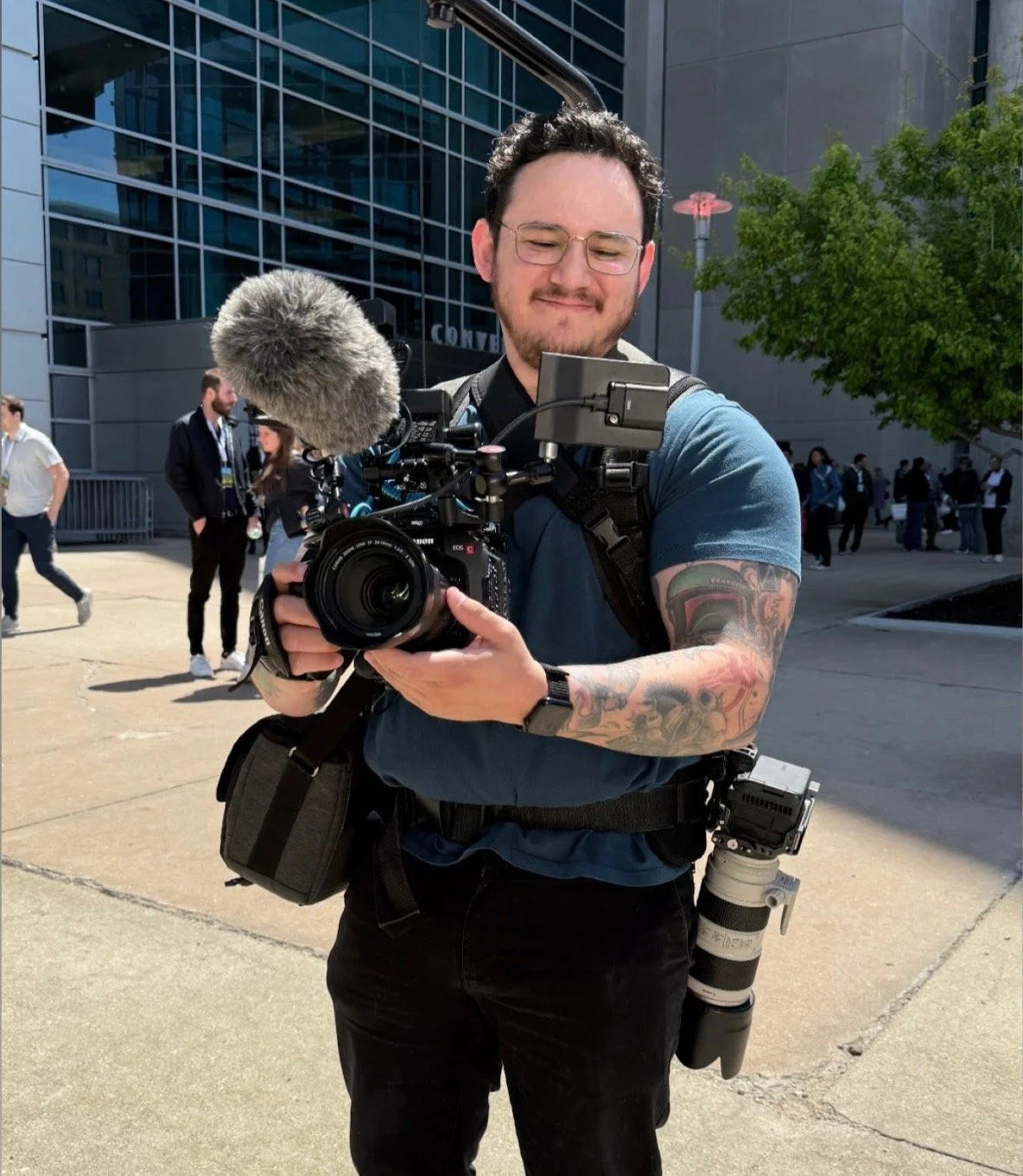 A man holding a professional video camera with a microphone outdoors in an urban area, smiling, with several people and a modern glass building in the background.