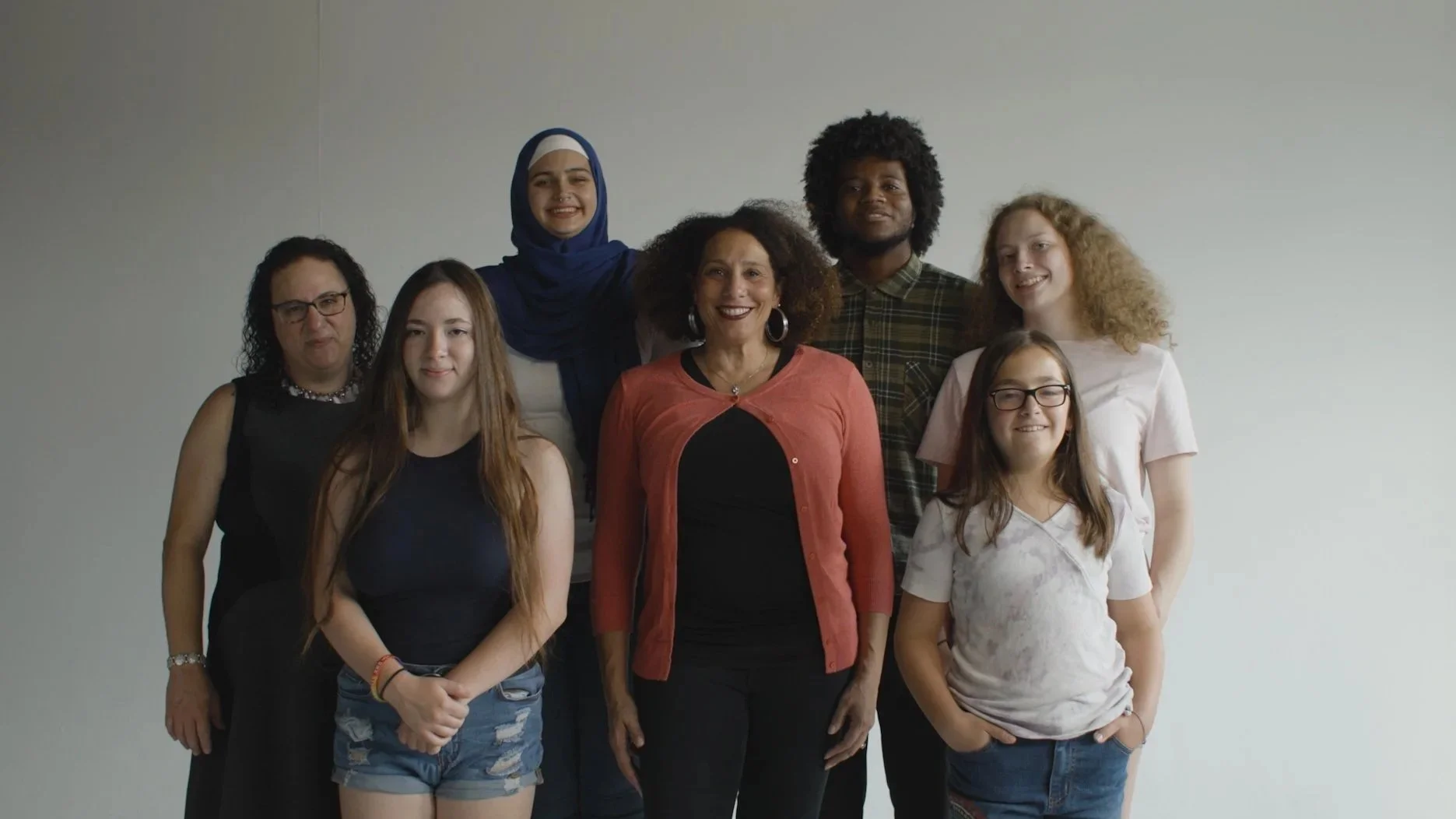 Group of diverse women and girls standing together in front of a plain white wall, smiling at the camera.
