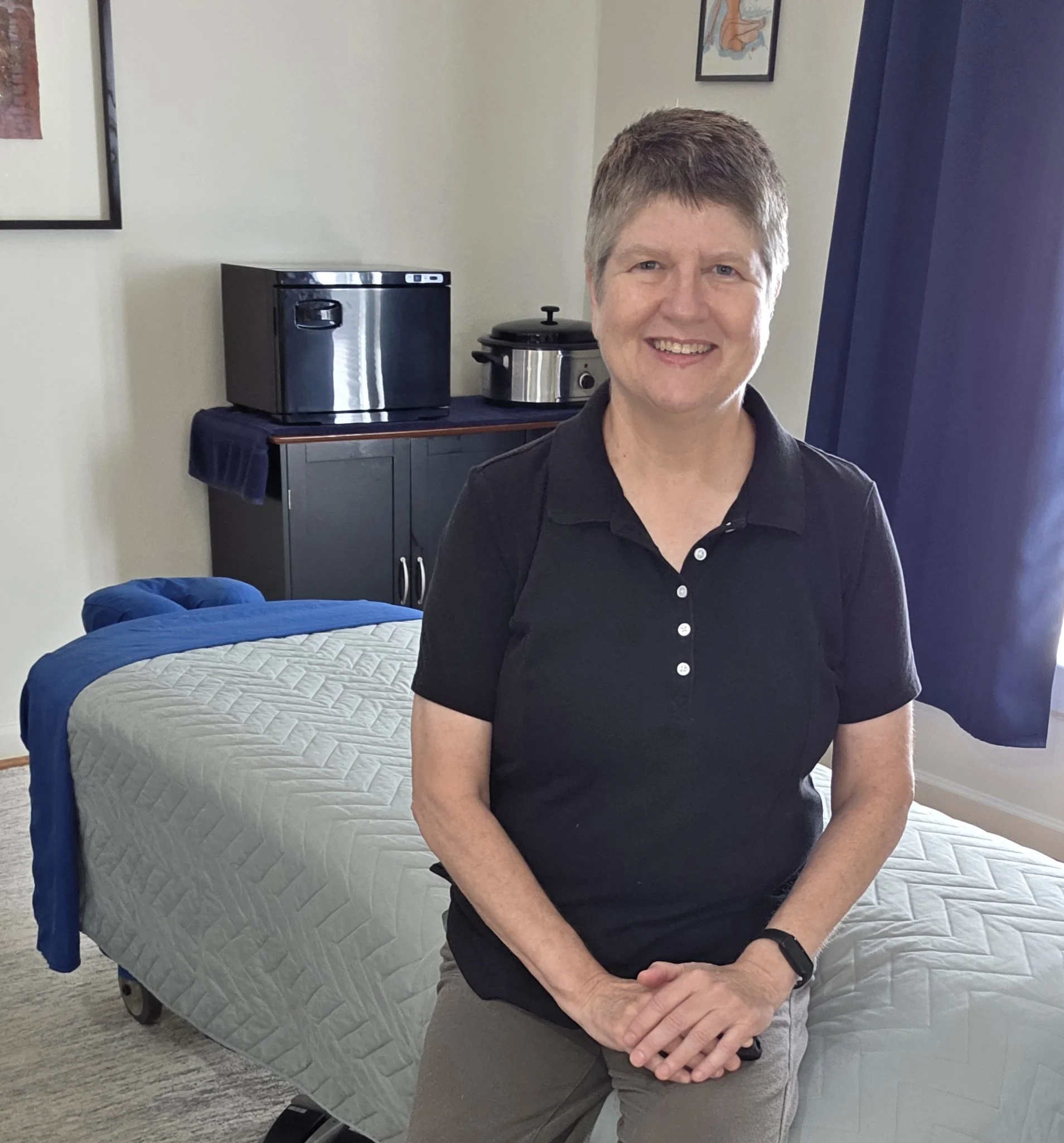 A woman with short hair smiling, standing in a room with a bed, a black mini fridge, and a slow cooker on a table behind her.