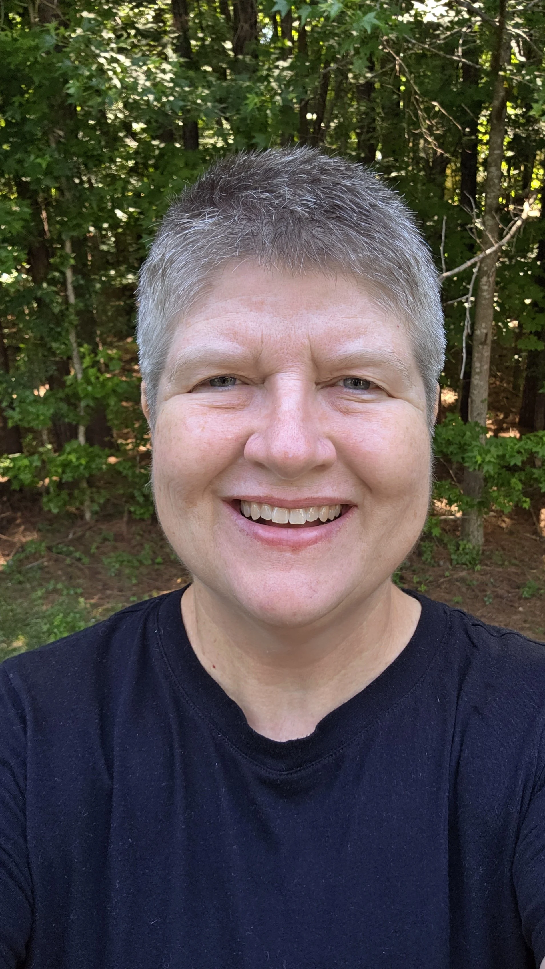 A smiling person with short gray hair, wearing a black shirt, standing outdoors in front of green trees.