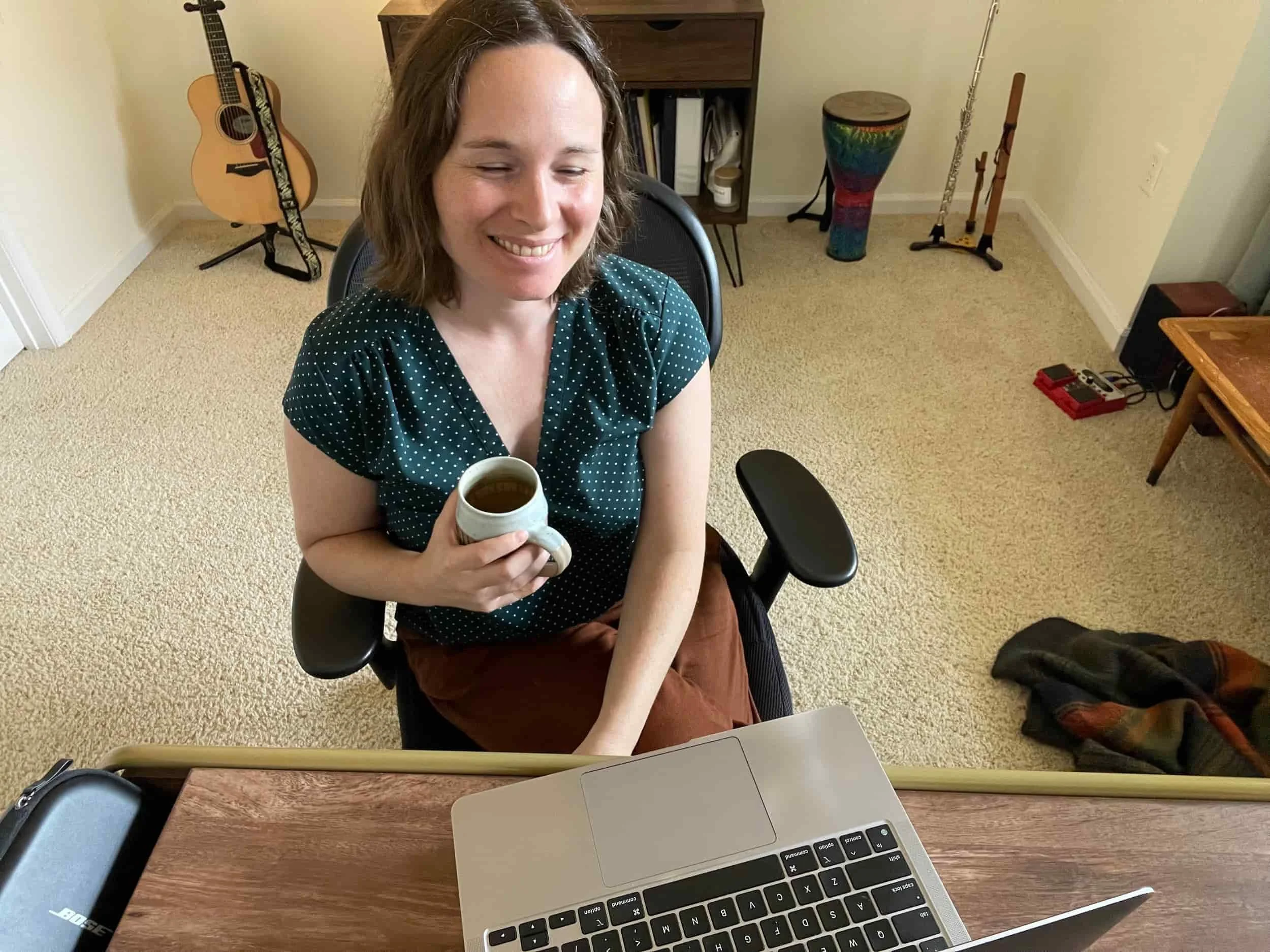 Woman smiling, holding a mug, sits at a desk with a laptop; musical instruments in the background.
