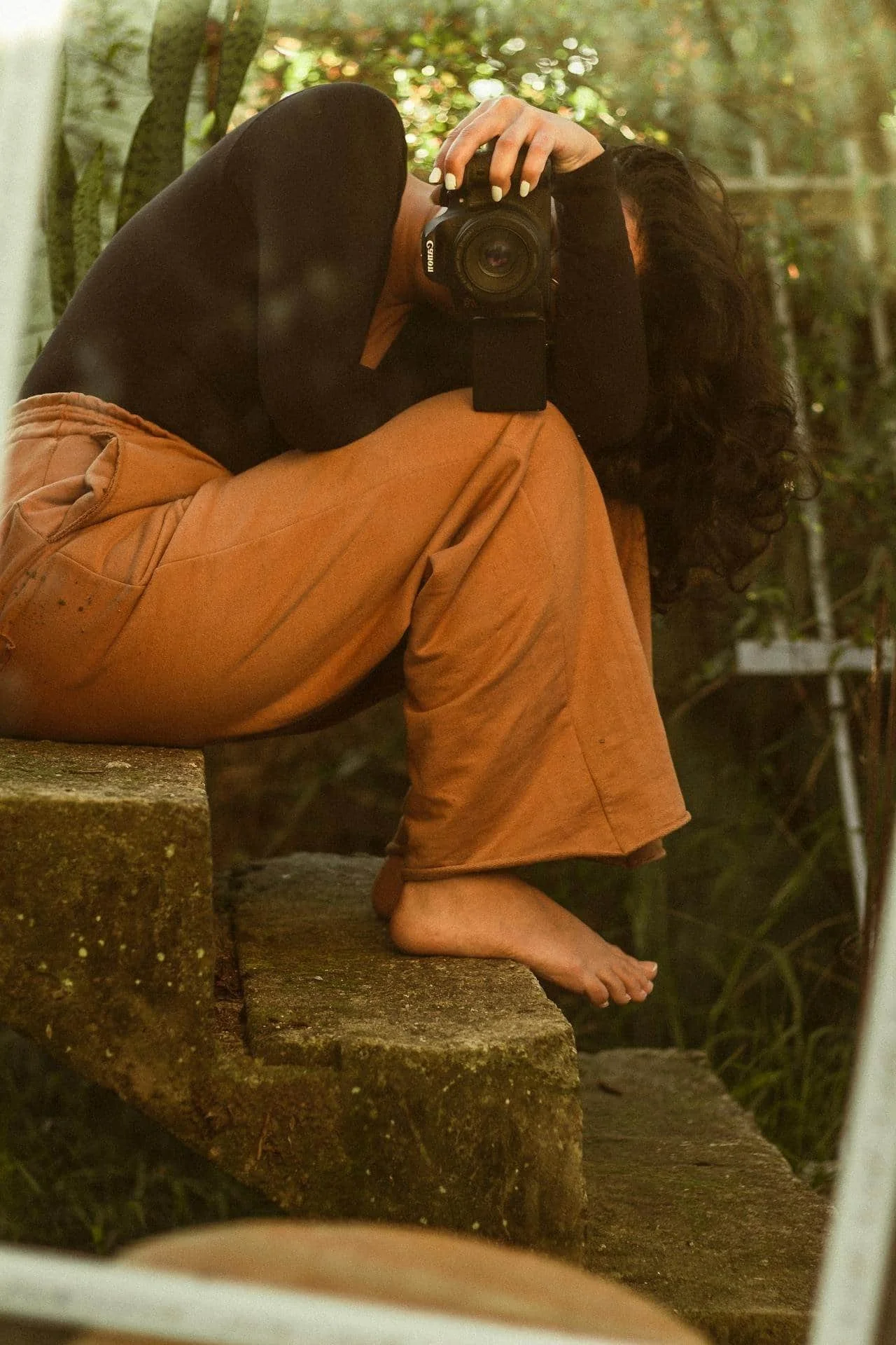 A person sitting barefoot on stone steps, wearing brown pants and a black top, taking a photo with a camera.