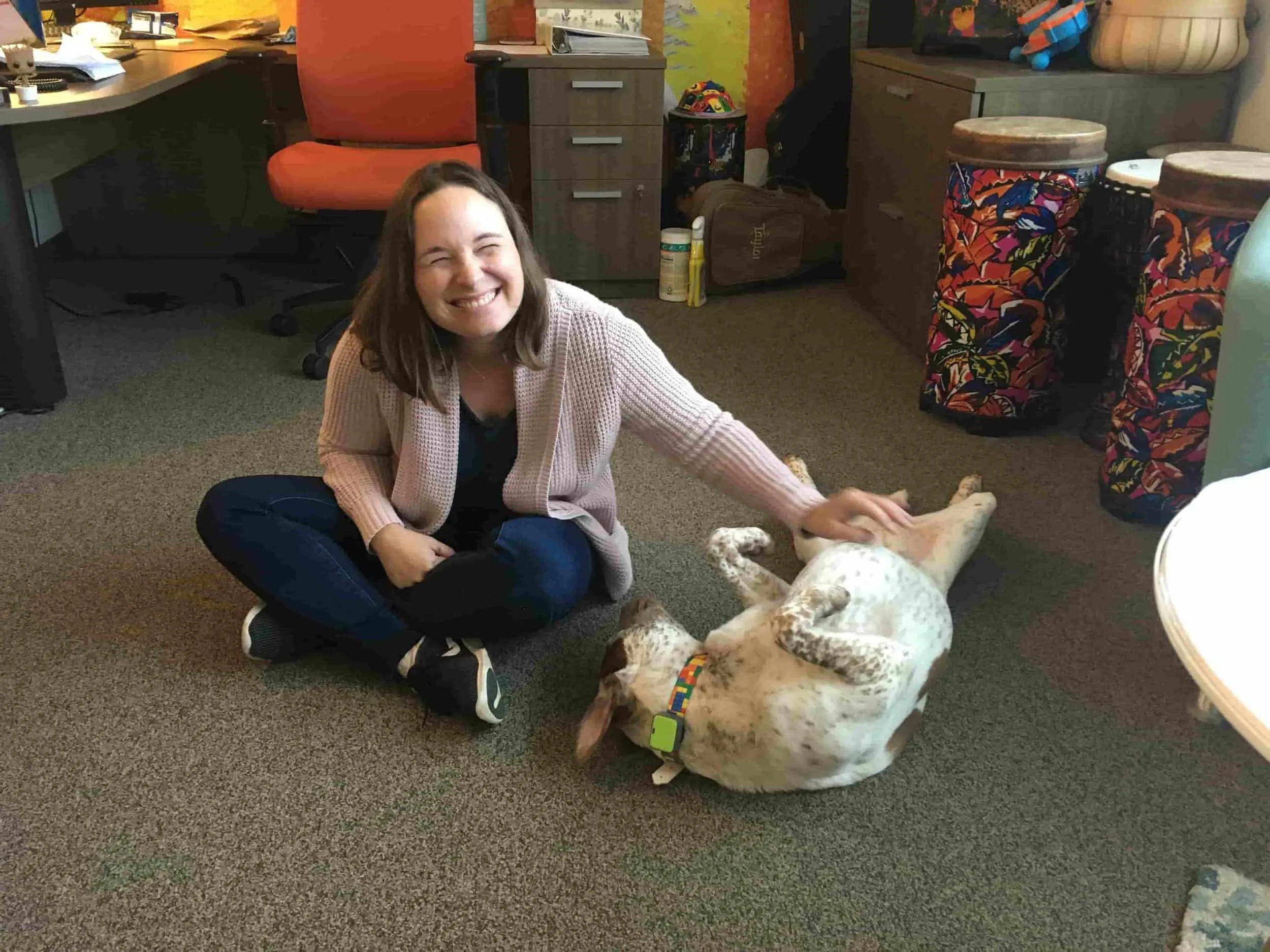 Smiling woman sitting on the floor, petting a happy dog lying on its back in an office with colorful instruments and decor.