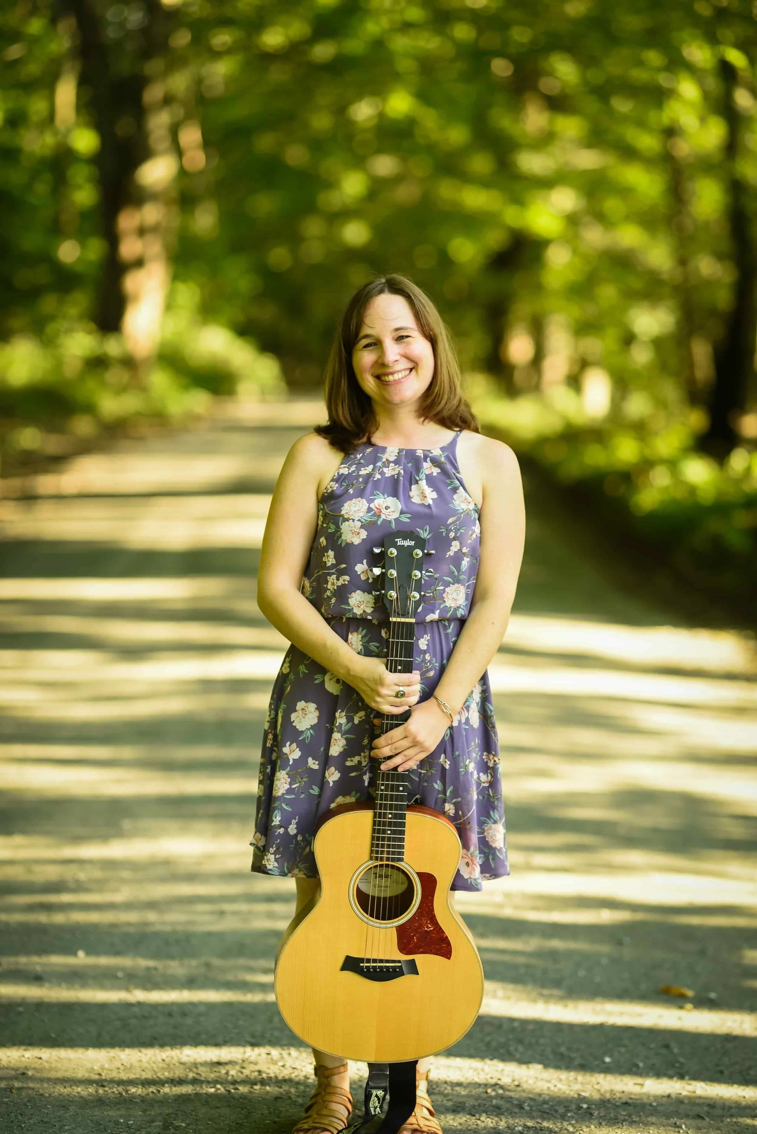 Woman in a floral dress stands on a sunlit forest path, smiling and holding an acoustic guitar upright in front of her.
