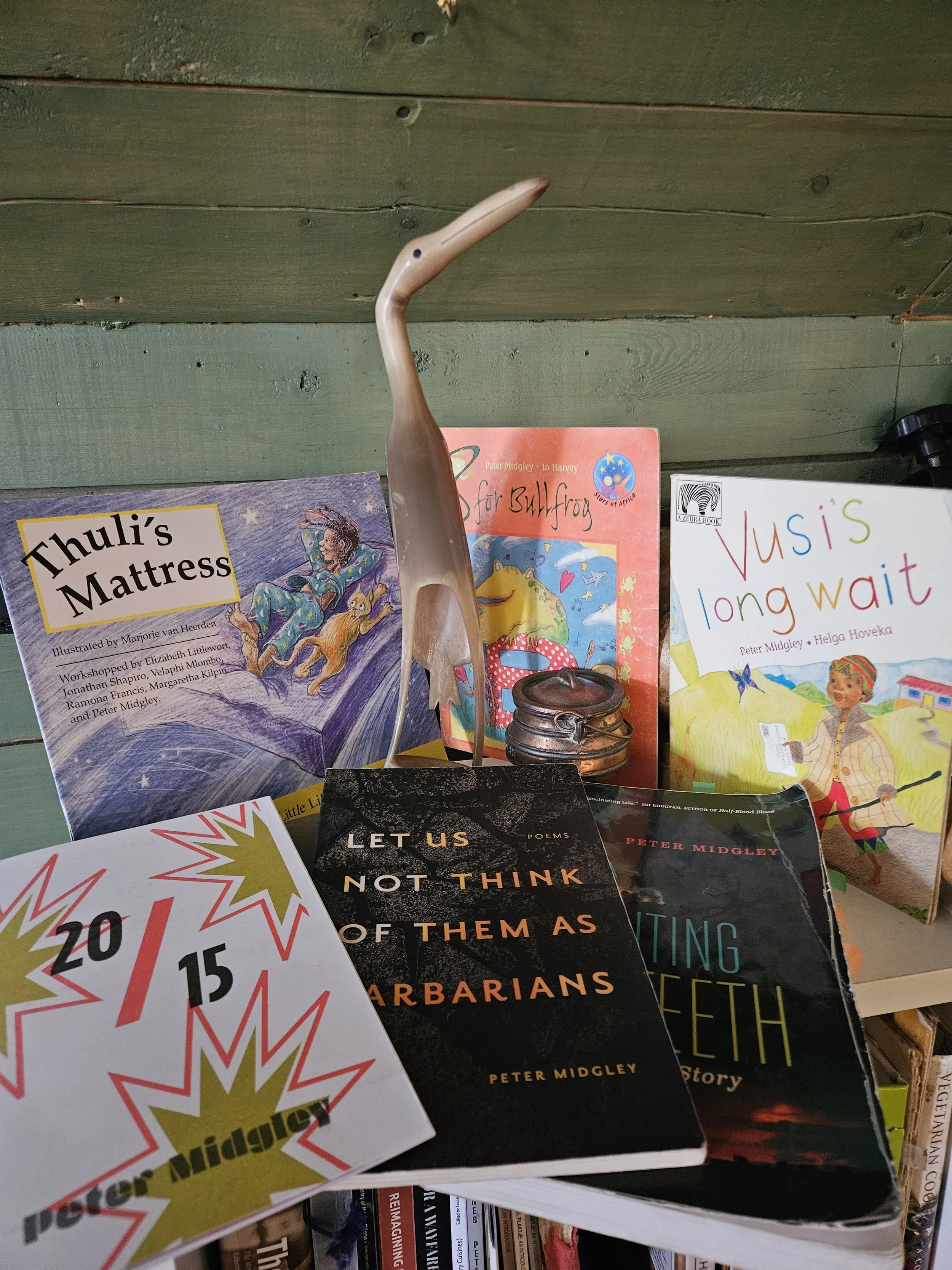 Six books on a table. with a bird figurine in the centre. In front of green-tinted wood. Three childrens books with colourful covers standing on table. Three paperbooks lay down in front of the other three. The 20/15 chapbook is bottom left.