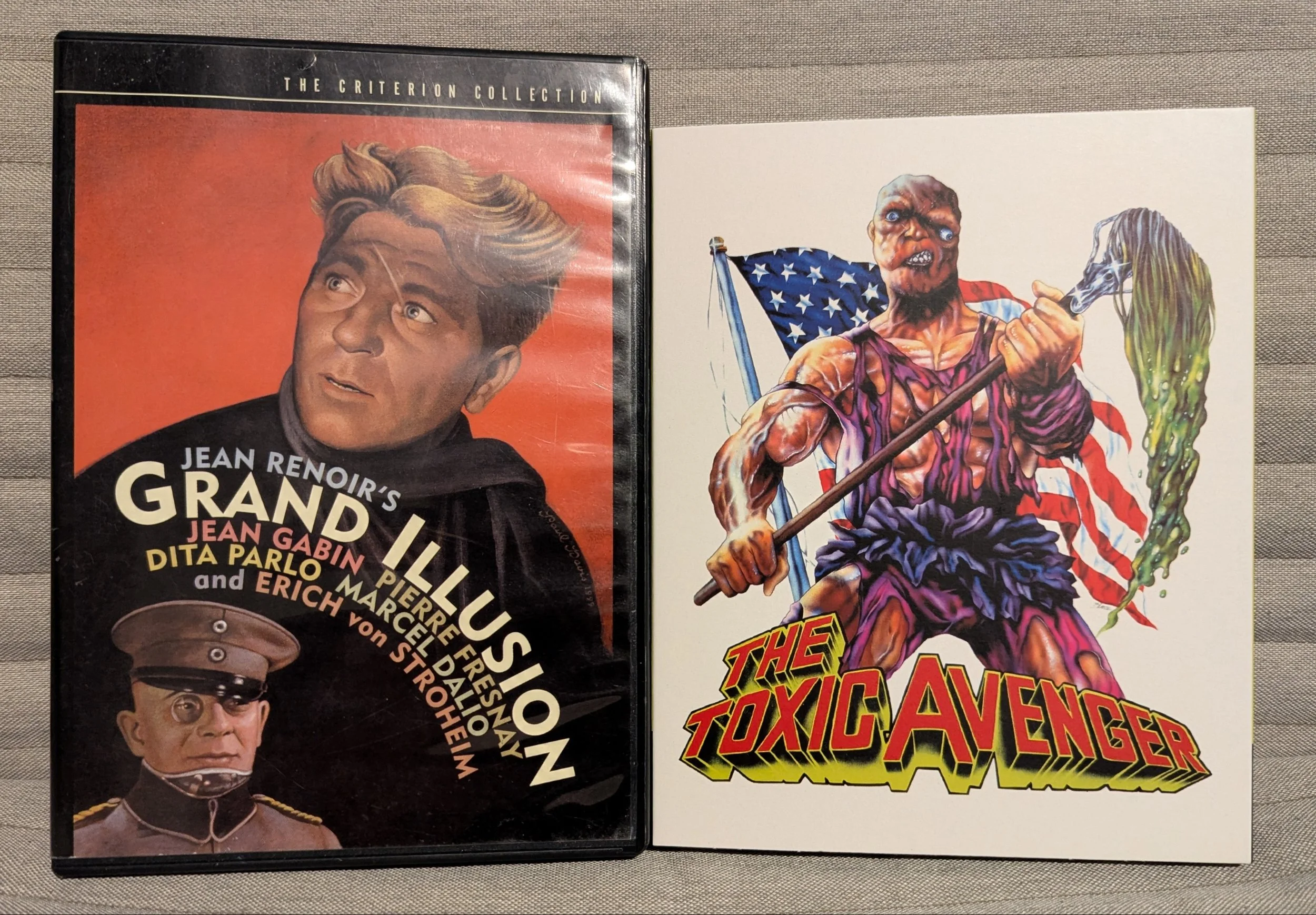A DVD and Blu-ray case beside each other. Left is "Grand Illusion." Two men illustrated realistically on the cover, red background. Second movie is Toxic Avenger. White case with an animated mutated person holding a mop with green goo.