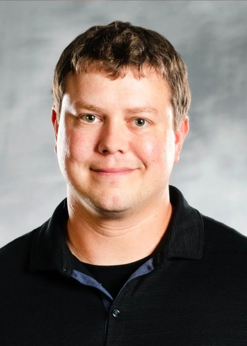 A headshot of a clean-shaven man with white skin smiles as he looks directly at the camera. e has light green eyes and short brown hair. He wears a black shirt with a collar. He is in front of a non-descript gray background.