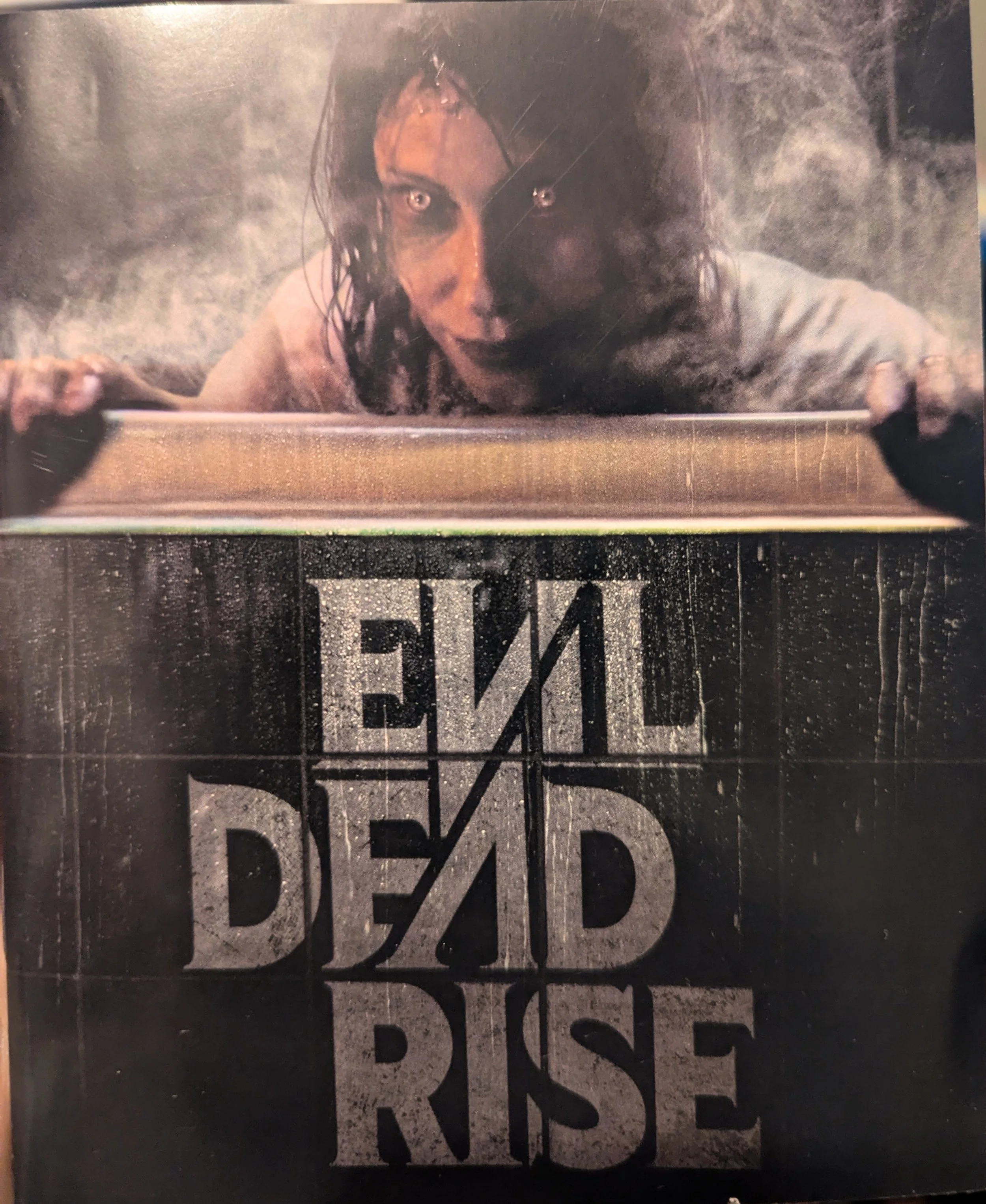 "Evil Dead Rise" booklet cover. The title of the film is on the bottom half over black background. The top half features a deadite woman from the film with smoke around her head.