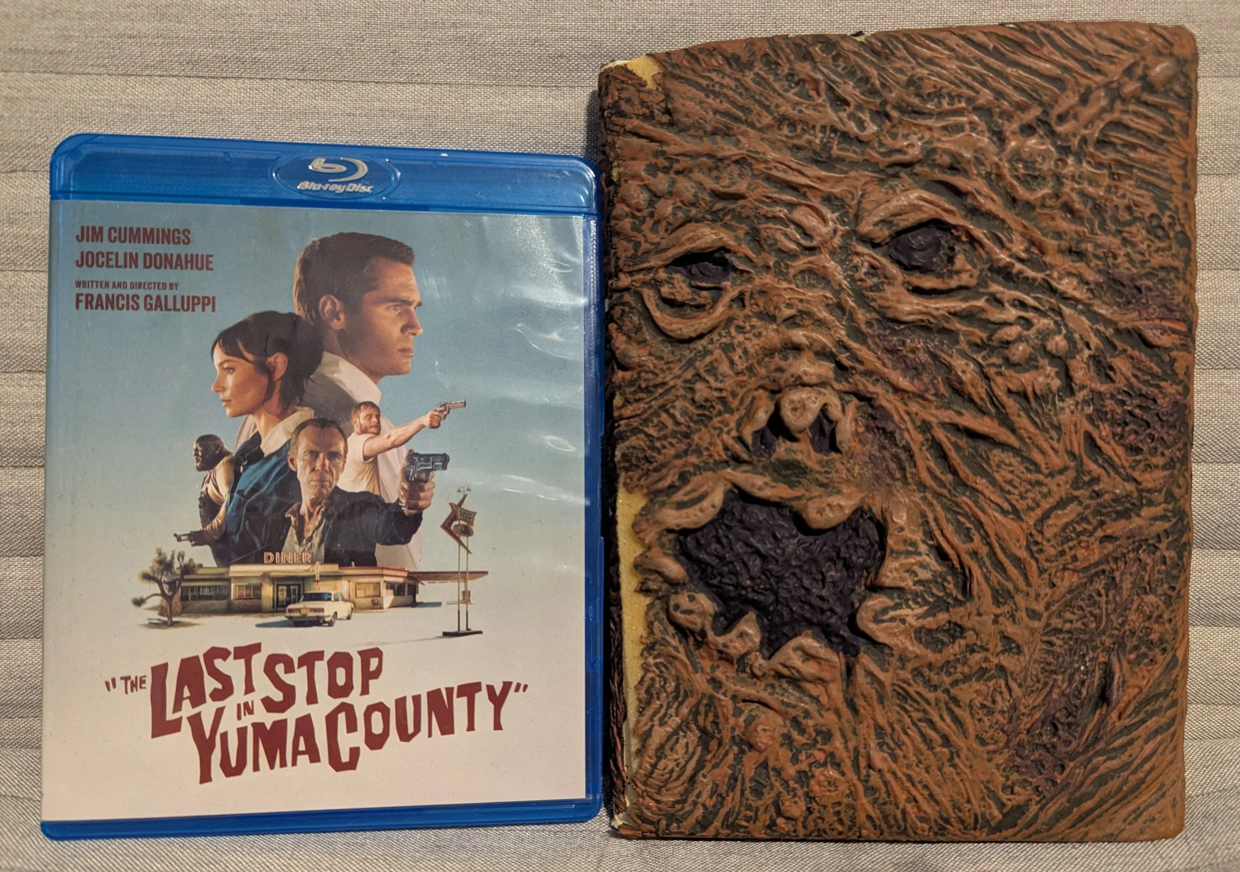 Two movie cases. On left is Last Stop in Yuma County, movie title on case and a collage of faces over diner on blue back. On right is replica of Necromonicon book from The Evil Dead, wrinkly brown face with mouth and eye holes.