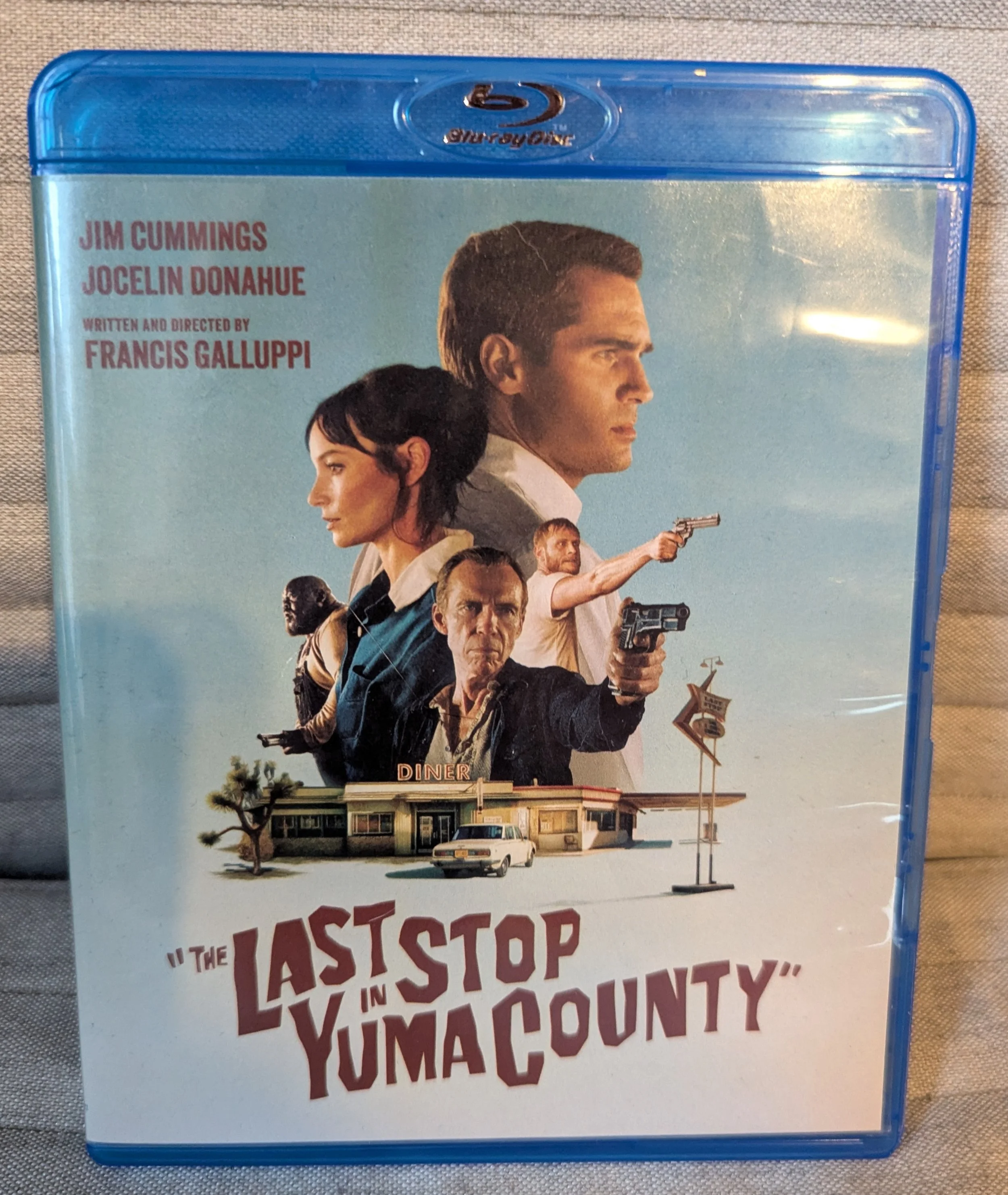 The Blu-ray case for "The Last Stop in Yuma County." The movie title is on the cover at the bottom. Above the title is collage of five characters from the movie over an image of a diner with a white car in front. Cover has light blue background.