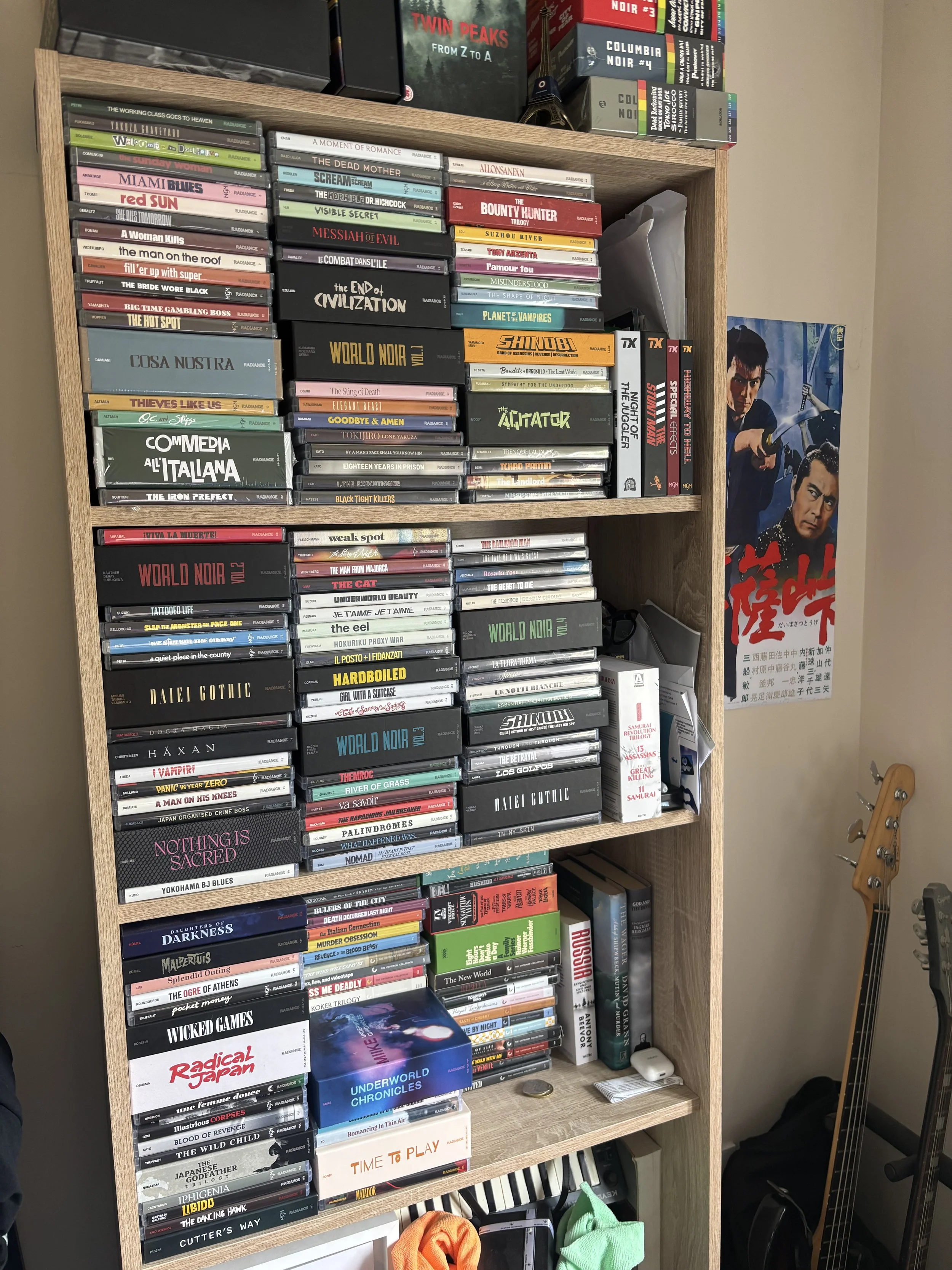 A bookshelf with four shelves that are full with Blu-rays stacked horizontally. Most are Radiance Films releases, which are various colours with the movie's title. The top of the bookshelf also has boxsets on it. Tops of a bass and a guitar visible.