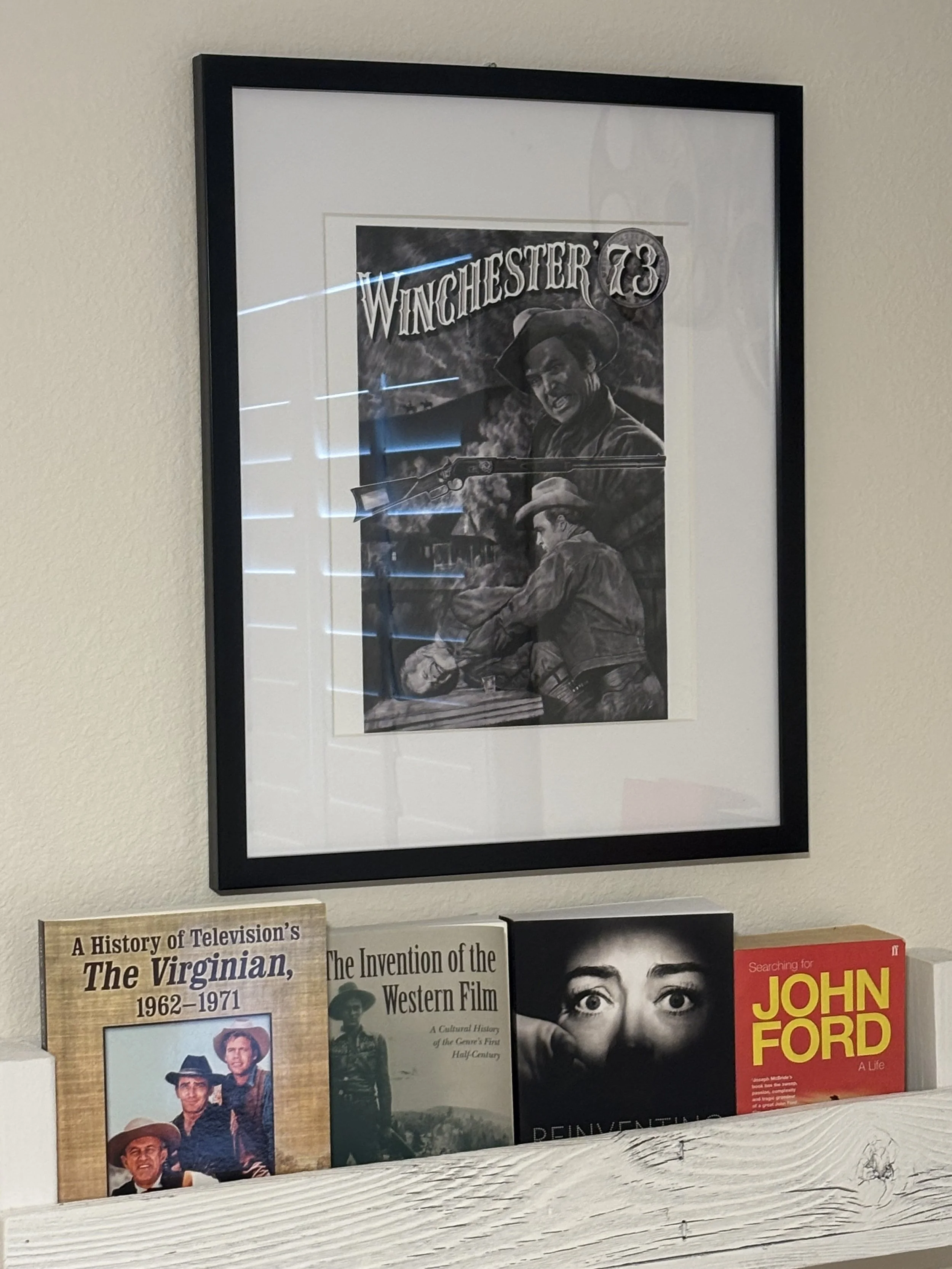 A poster framed hanging on a wall over four large books that stand upright on a white wood shelf. The poster is for Winchester '73. It is black and white with painted cowboys. The books are all Western-focused, including a red book on John Ford.