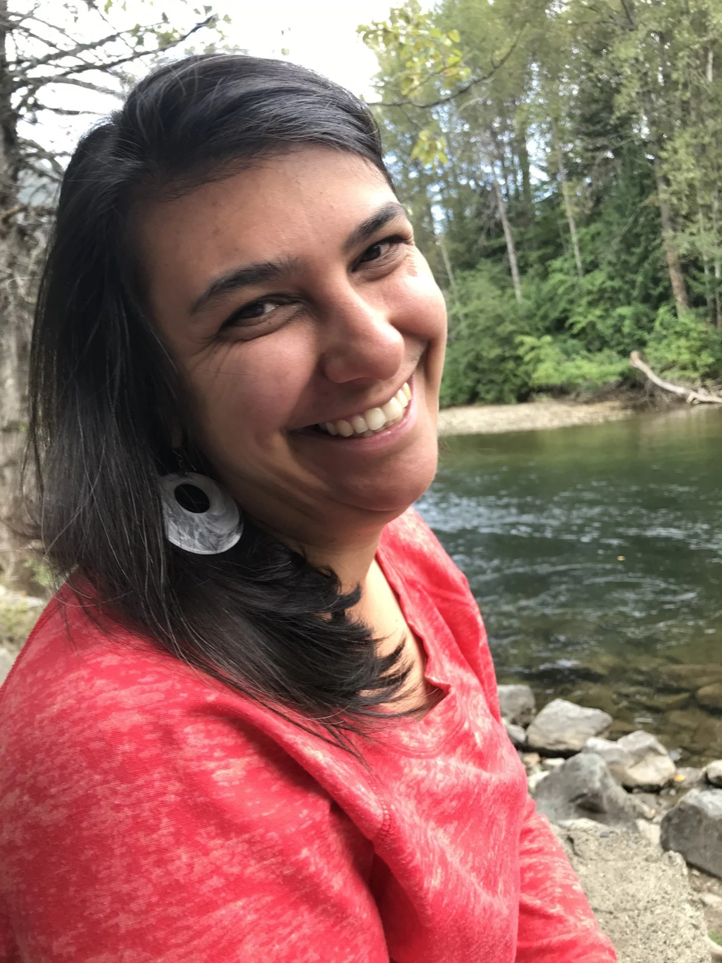 A close-up image of a woman smiling directly into the camera with her head turned. She is in front of a river with tall green trees on the other side. She wears a red light sweater and a gray earring, and she has brown hair and brown eyes.