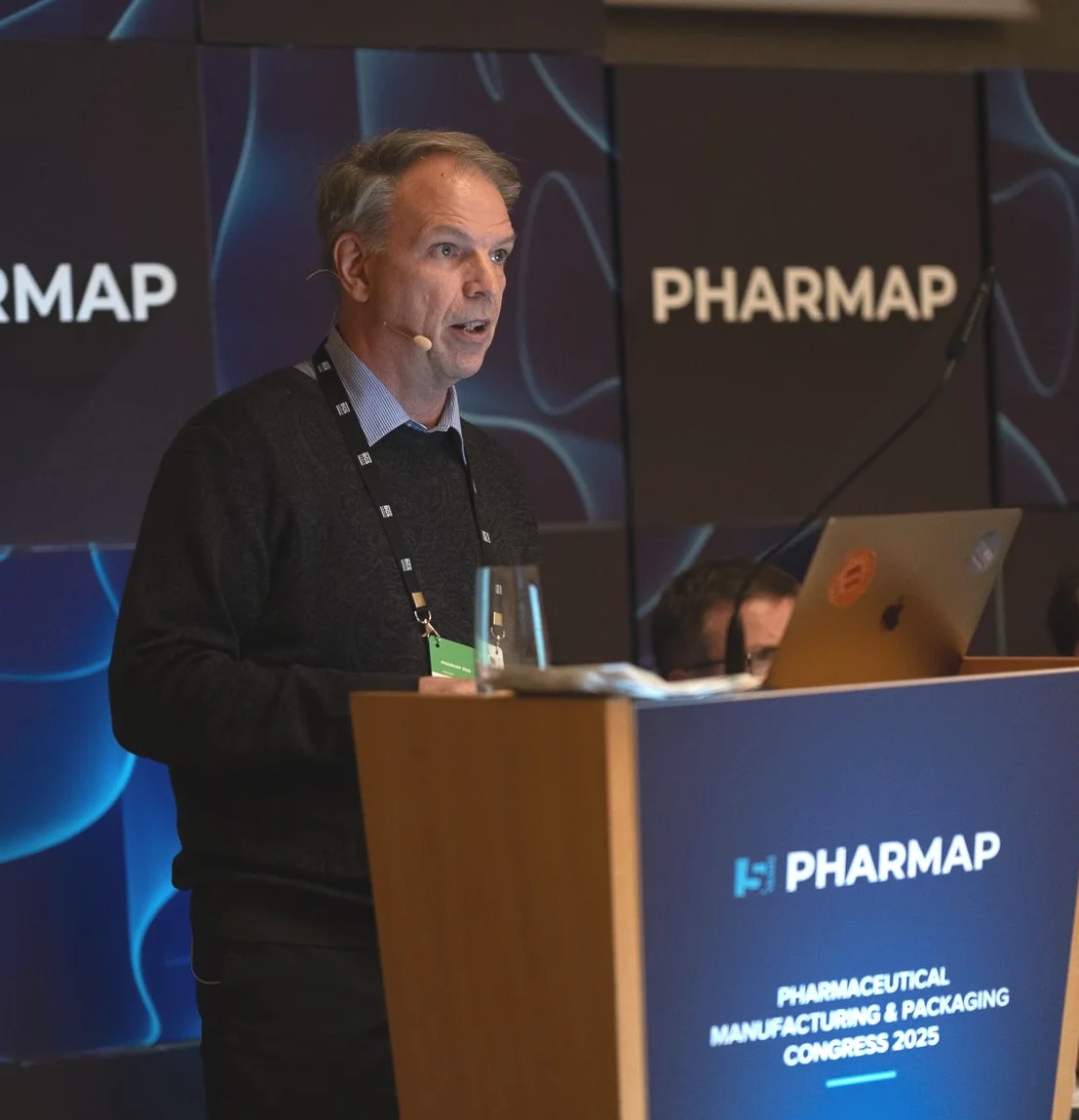 A man standing behind a podium in front of a blue screen with the word "pharmap" on it twice. He is a white-skinned male with gray hair, mouth open mid-speech. He wears a navy sweater with a blue collar and a tag on a lanyard around his neck.