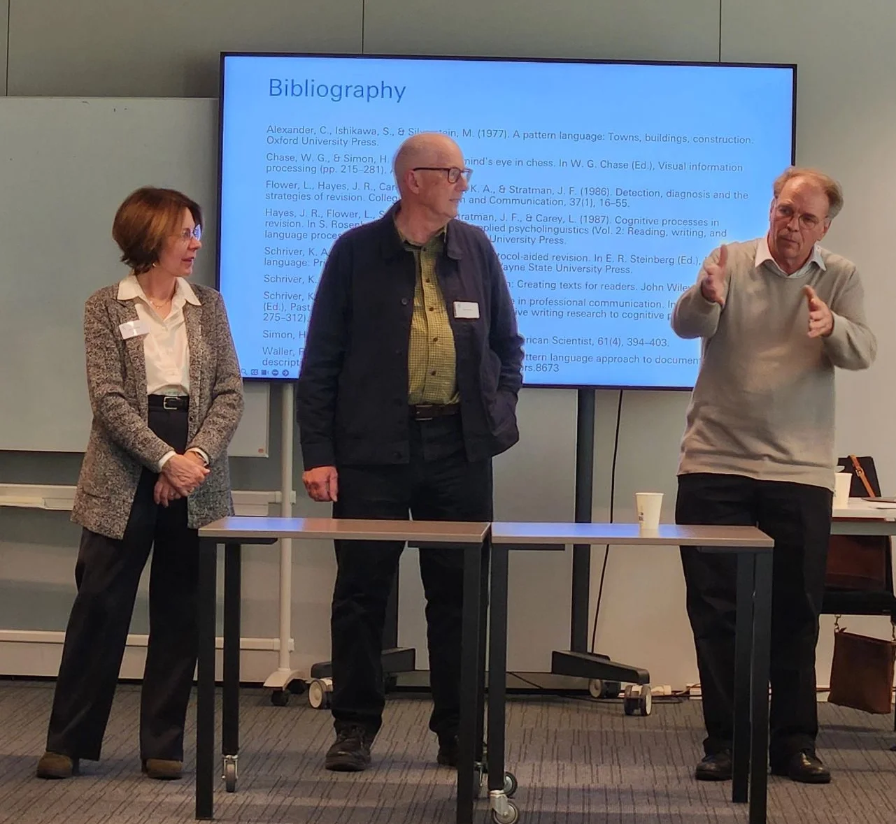 Three people (with Karel on the right) stand on stage with a screen behind them displaying a bibliography. The two other people, a woman with brown hair and white skin and a bald man with white skin, look at Karel as he speaks. Two tables in front.