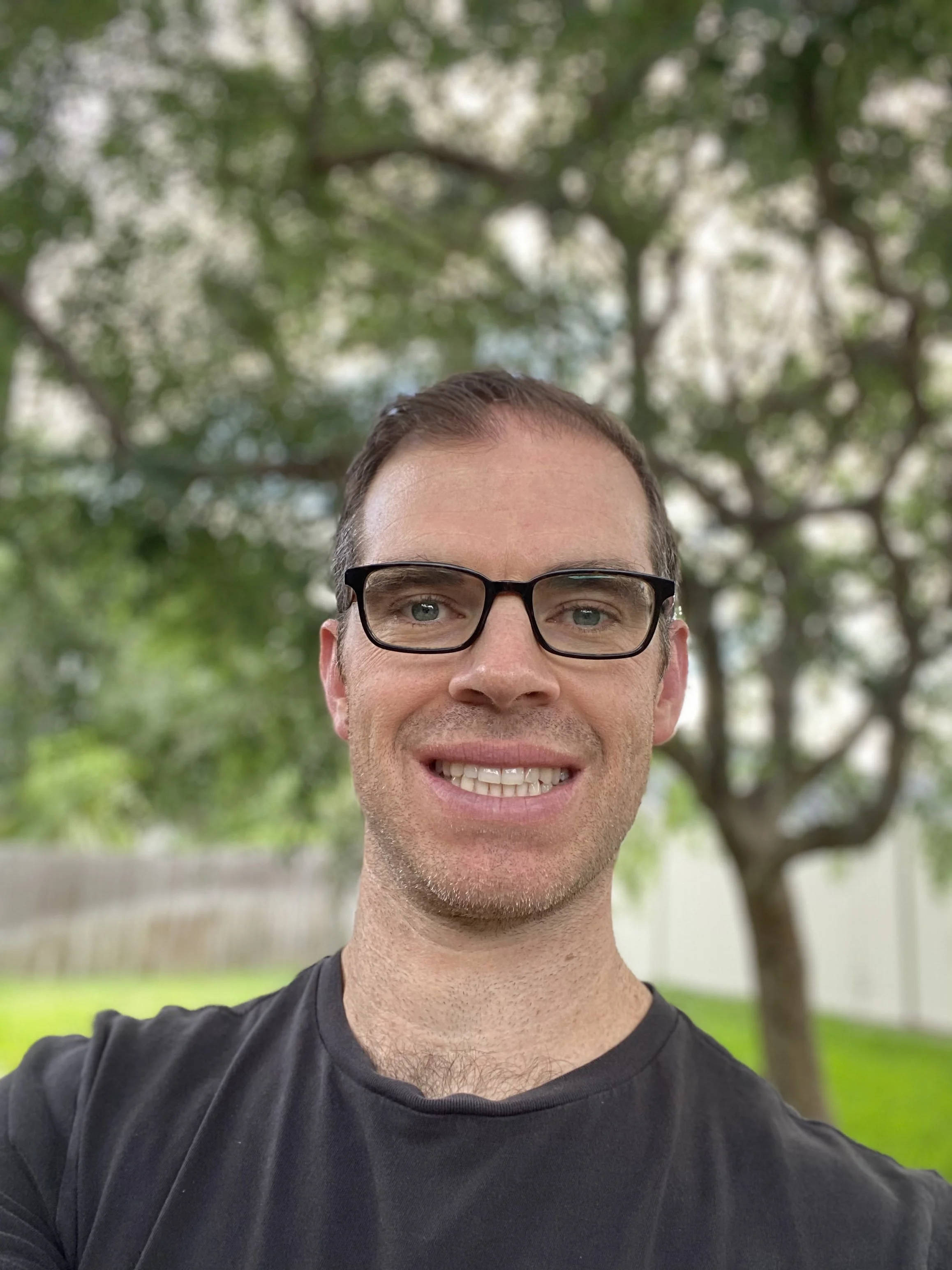 A selfie of a man smiling. He has short brown hair, thin glasses with black frames, light stubble, white skin, and wears a gray T-shirt, visible from the collar up. He is outside with trees behind him, though the background is blurred out.