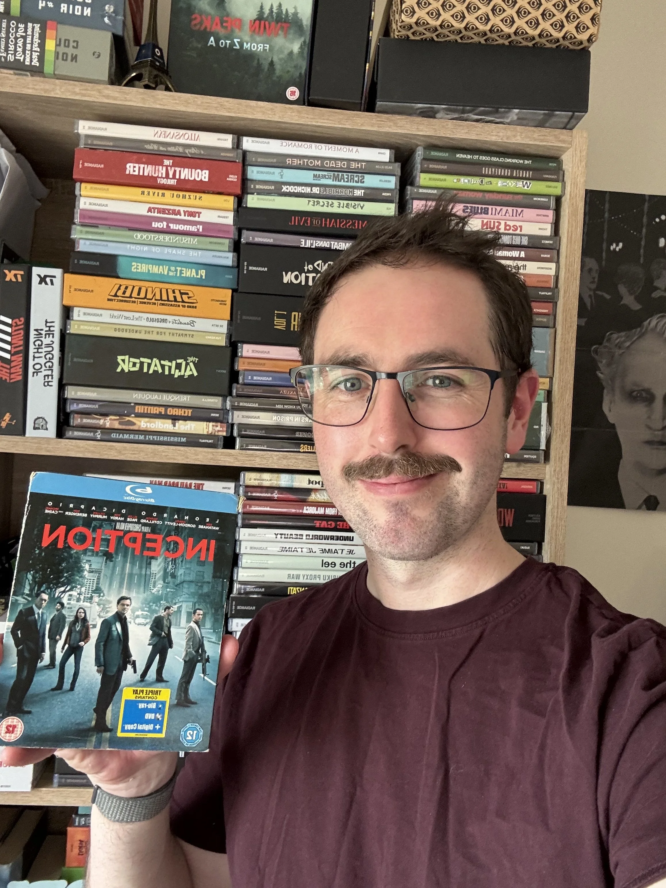A man with white skin, short brown hair, a moustache and glasses wearing a burgundy T-shirt smiles as he holds an Inception Blu-ray.. He is in front of a shelf with Radiance Blu-ray releases. Black and white poster beside shelf of men's faces.