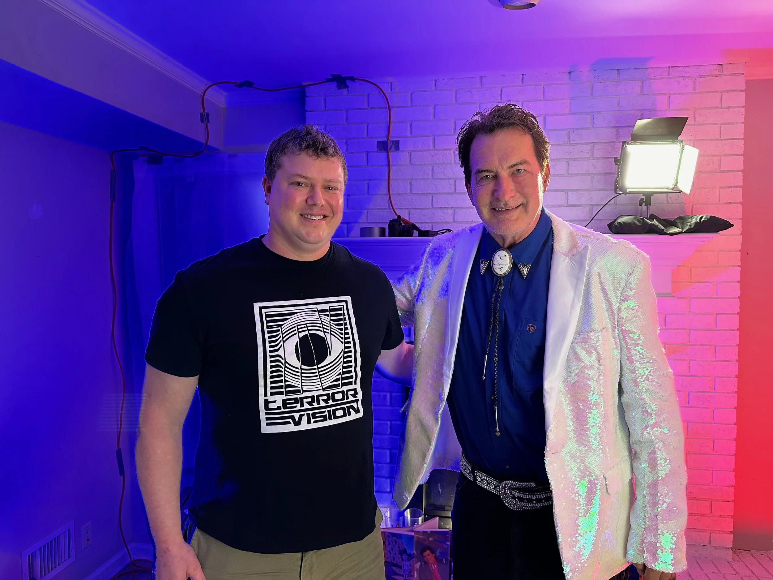 Two white men stand beside each other, Dodson on the left, Briggs on the right. Dodson wearing a black T-shirt with Terror Vision logo. Briggs wears shiny white jacket, blue dress shirt, neck piece, and bedazzled belt. Blue and pink lighting behind.